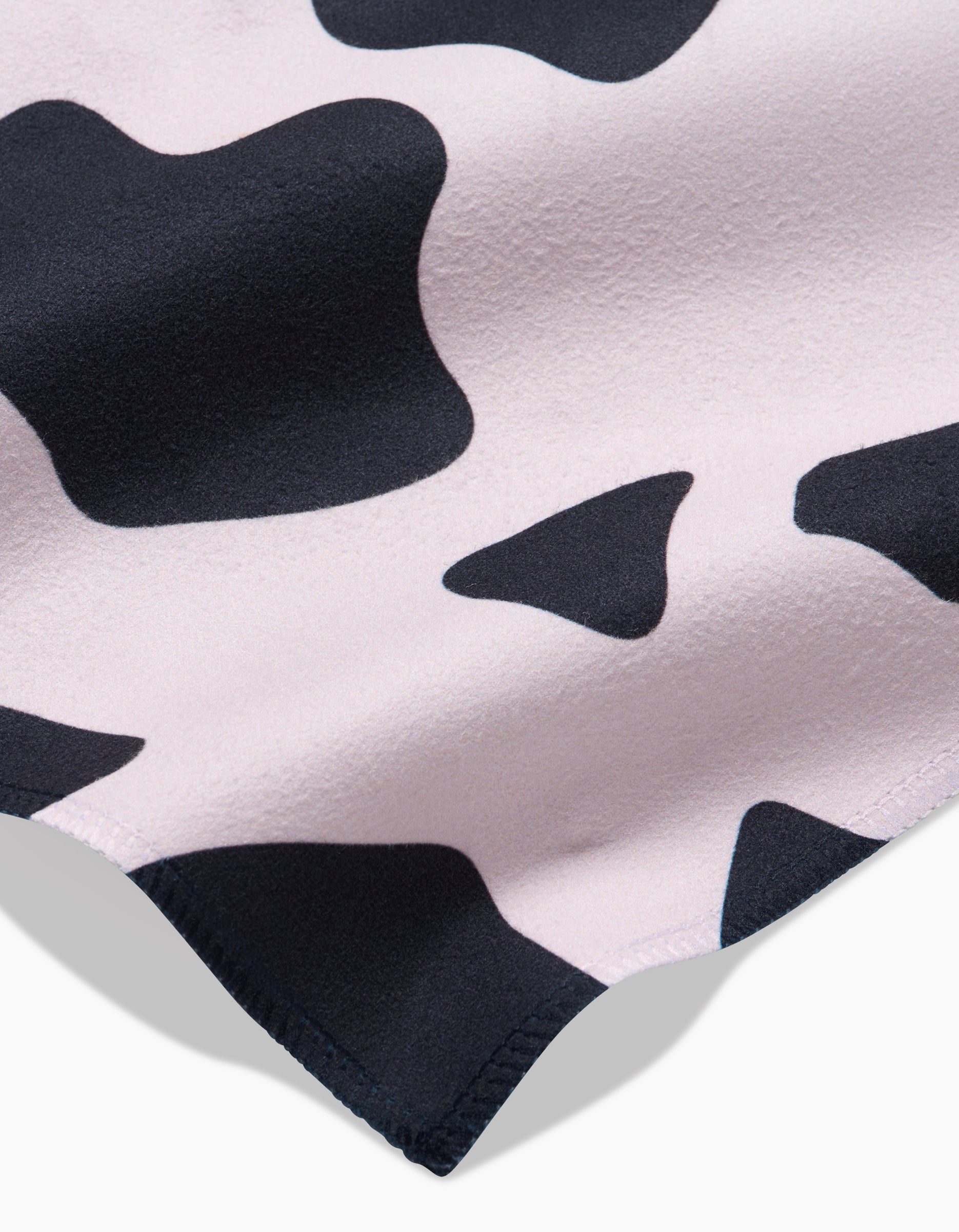 Close-up of Cow Print Black by GEOMETRY, a soft Geosuede fabric with irregular black spots on a light background and a visible stitched edge. This eco-friendly material is ideal for your Dinner Napkin Set.