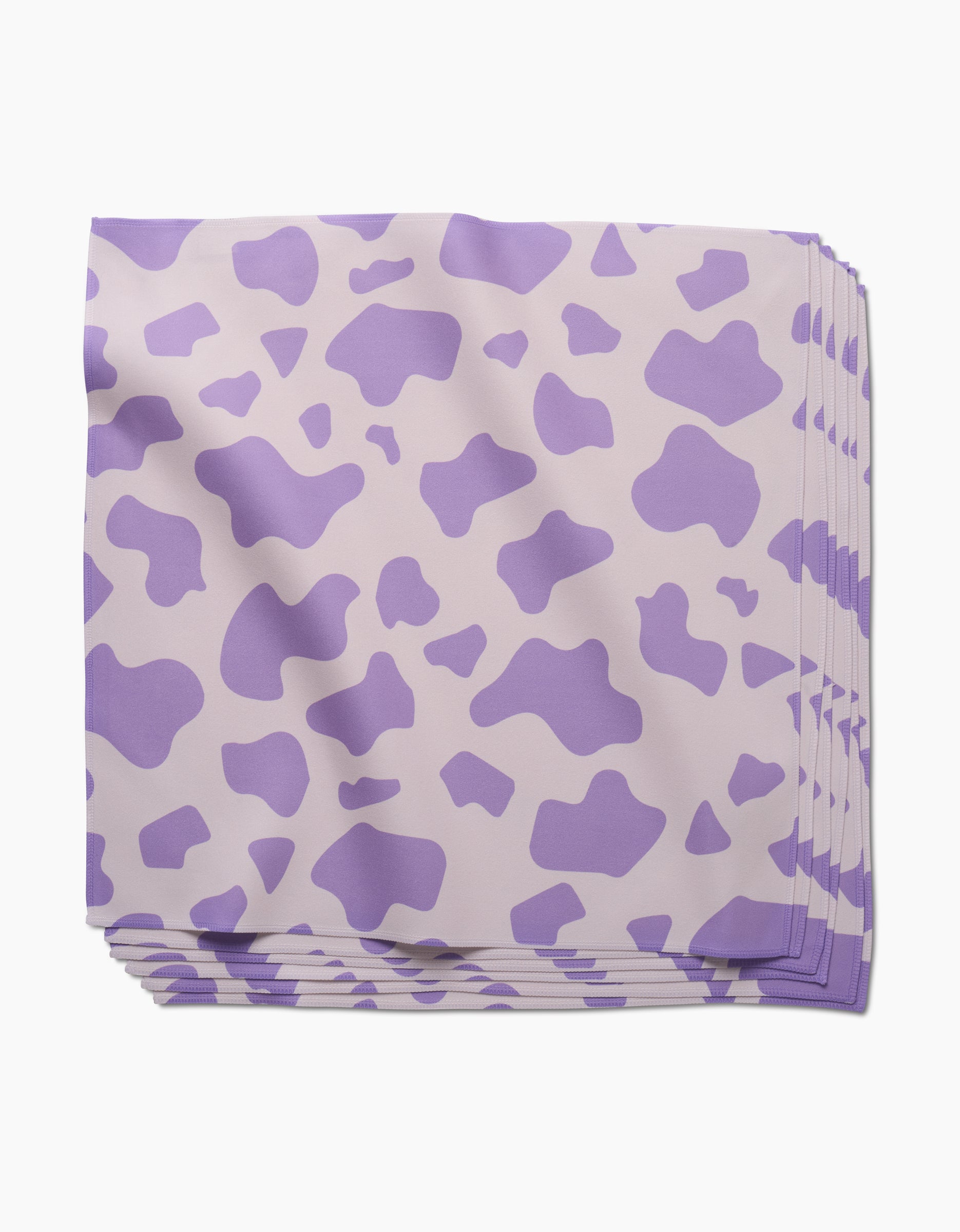 A neatly stacked set of GEOMETRY Cow Print Lilac absorbent cloth napkins featuring a light purple cow-print pattern on a pale background, displayed on a white surface.