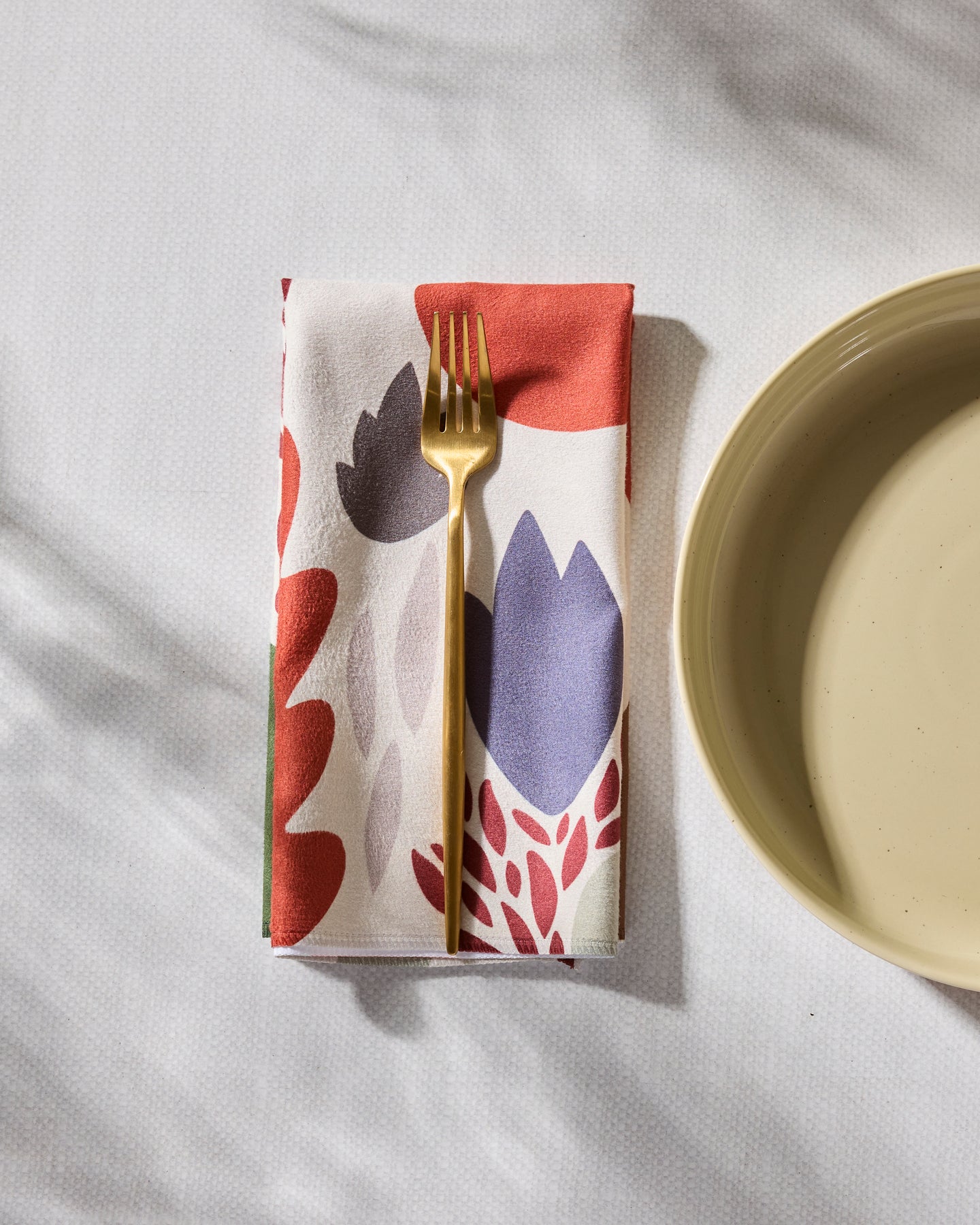 A gold fork rests on a Geometry Cut Paper Leaves Cream recycled dinner napkin, featuring a colorful floral pattern, beside a beige plate on a white textured surface.
