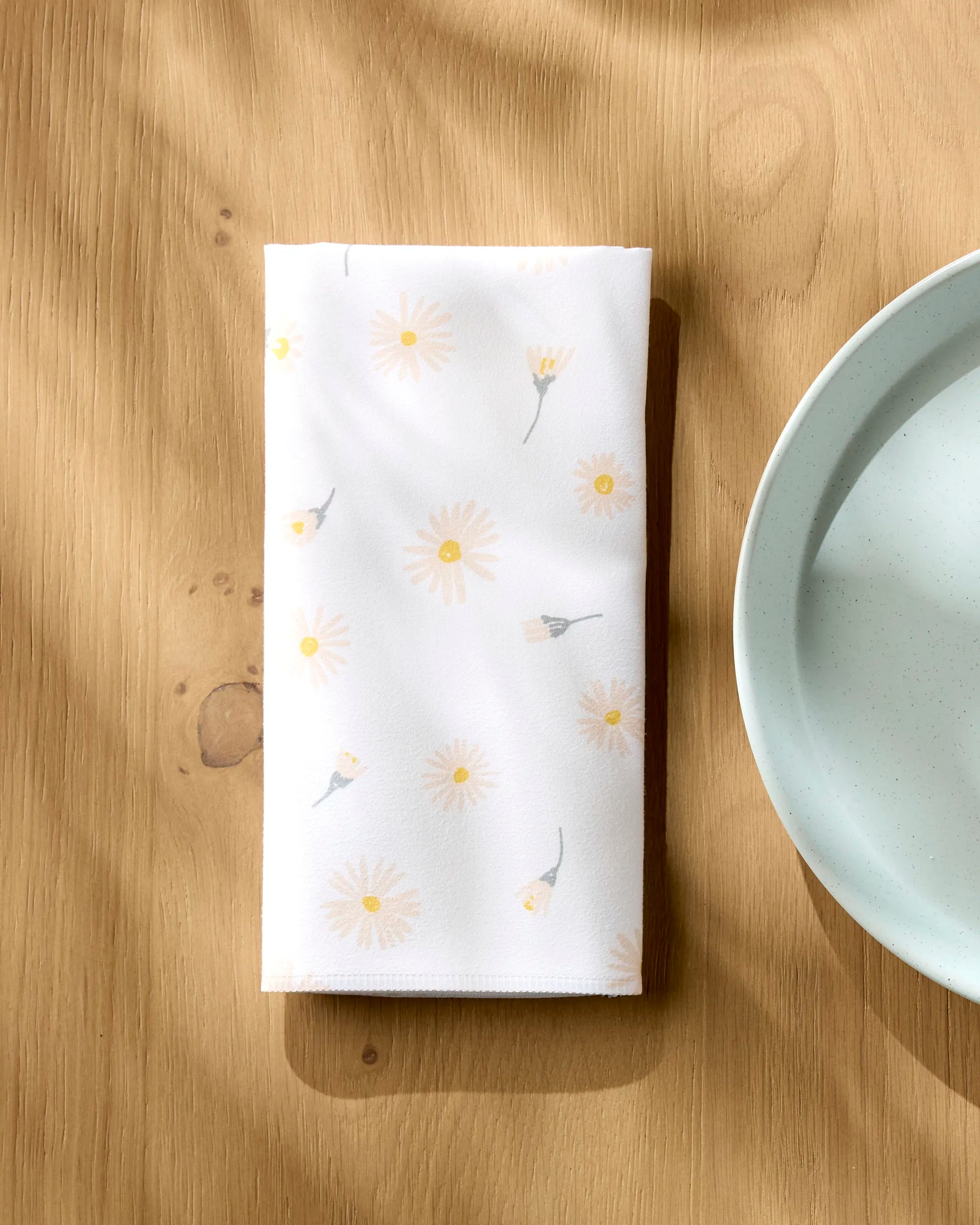 The Hufton Studio Daisy Dust reusable napkin, featuring a light yellow daisy pattern, is neatly folded on a wooden table beside a ceramic water bowl as sunlight casts soft shadows across the eco-friendly scene.