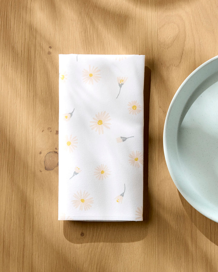 The Hufton Studio Daisy Dust reusable napkin, featuring a light yellow daisy pattern, is neatly folded on a wooden table beside a ceramic water bowl as sunlight casts soft shadows across the eco-friendly scene.