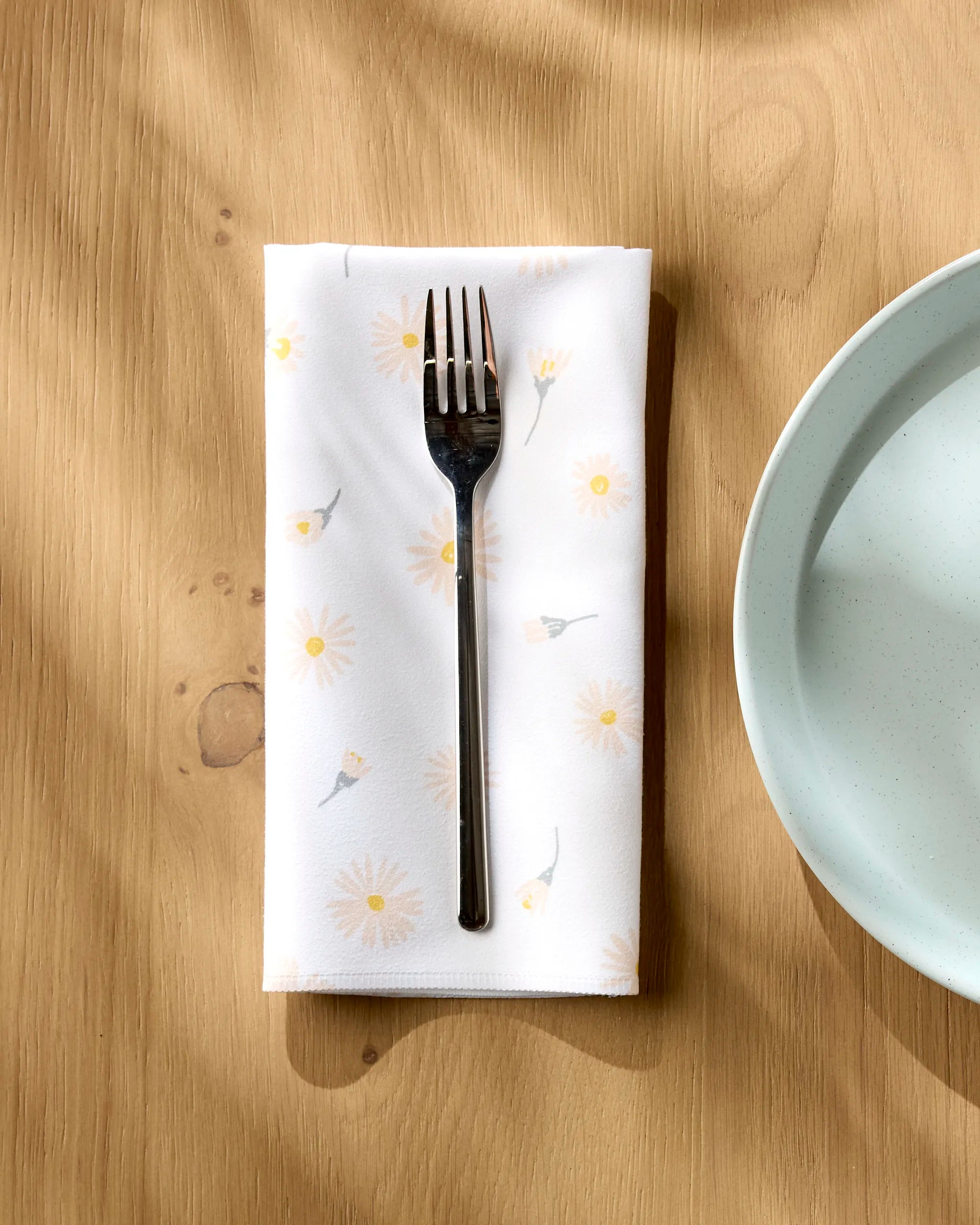 A Hufton Studio Daisy Dust metal fork rests on a recycled dinner napkin with a daisy pattern, placed beside a light blue ceramic plate on a wooden table as sunlight casts soft shadows across the scene.