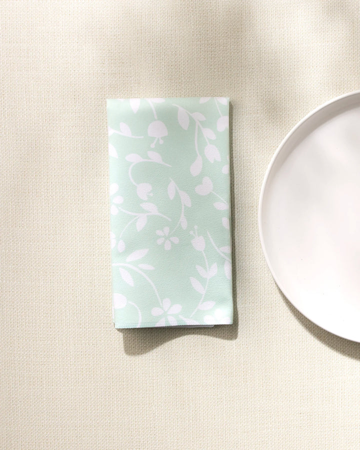 A folded GEOMETRY "Dandy" mint green dinner napkin set with a white floral design rests on a light textured surface next to a round white plate.