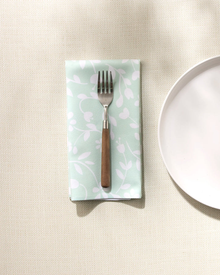 A GEOMETRY Dandy dinner napkin with a white leaf pattern is neatly folded on a light textured tablecloth beside a plain white plate, holding a fork with a wooden handle.