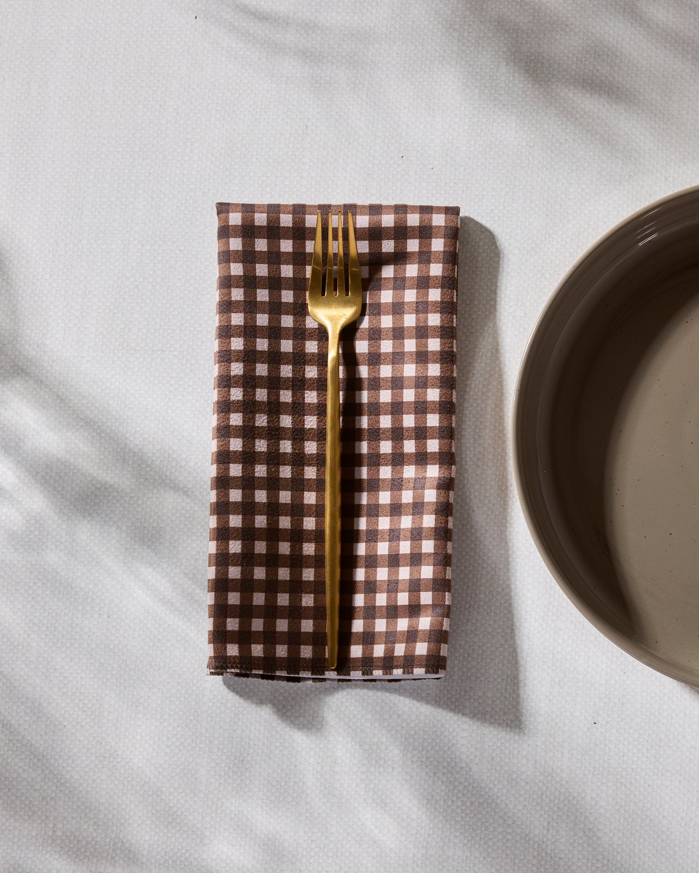 A gold fork rests on Geometry’s Deep Hearth brown and white checkered dinner napkin, folded beside a beige ceramic plate on a white tablecloth with soft shadows.