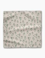 The Dirty Martini dinner napkin set by Rosa Kichline is light beige, made from recycled fabric, and showcases a repeating pattern of green martini glasses and green olives scattered across the surface.