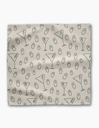 The Dirty Martini dinner napkin set by Rosa Kichline is light beige, made from recycled fabric, and showcases a repeating pattern of green martini glasses and green olives scattered across the surface.