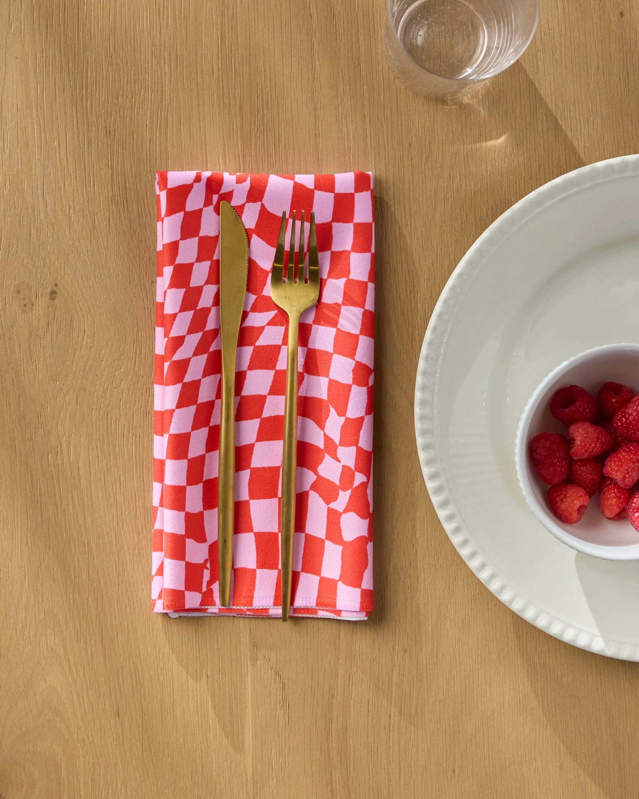 A gold knife and fork rest on Geometry’s Distorted Love red and white checkered reusable napkin beside a white plate, a bowl of strawberries, and a glass of water on a wooden table.