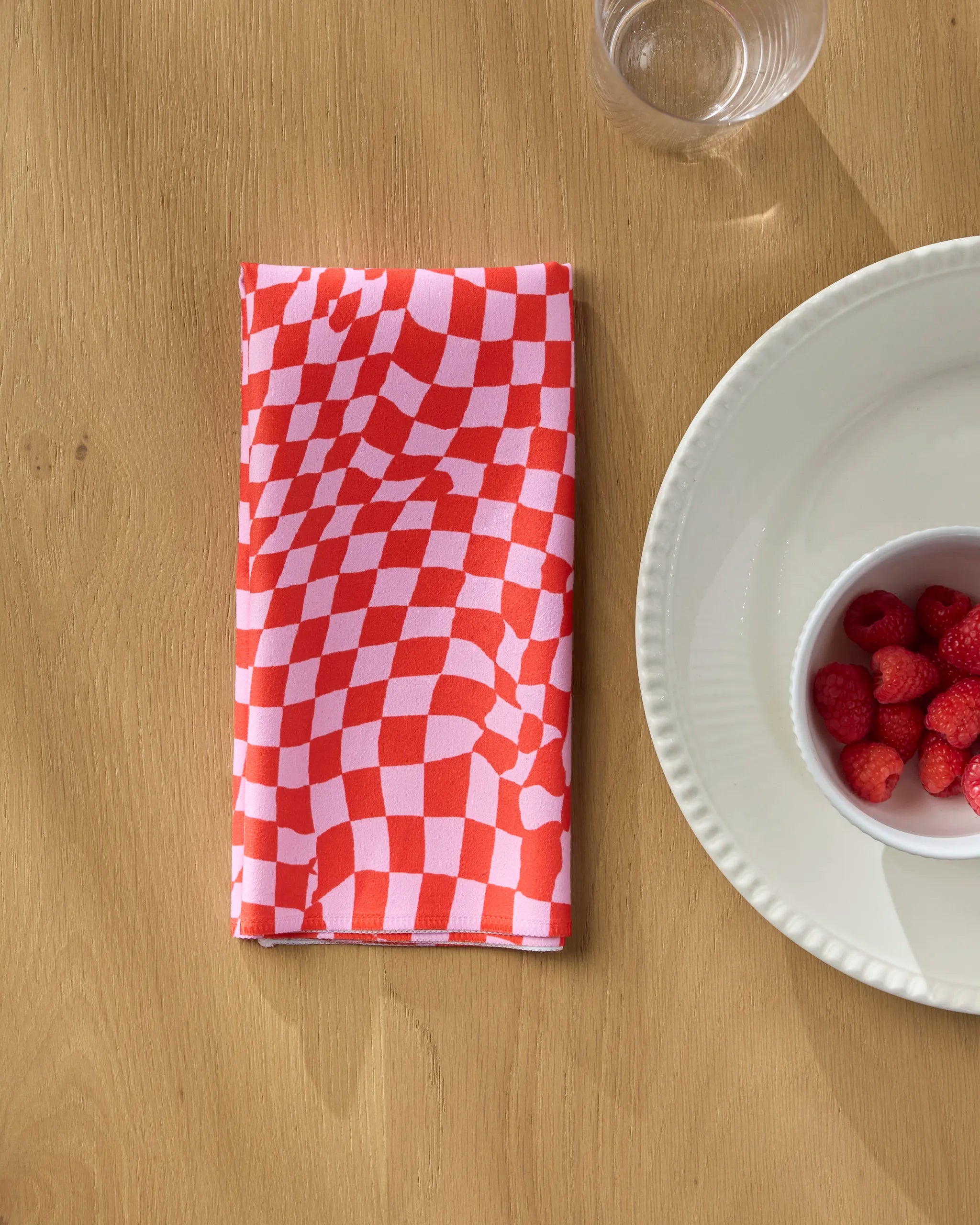 The Geometry "Distorted Love" red and white checkered reusable napkin is folded on a wooden table next to a clear glass, a white plate, and a bowl of raspberries.