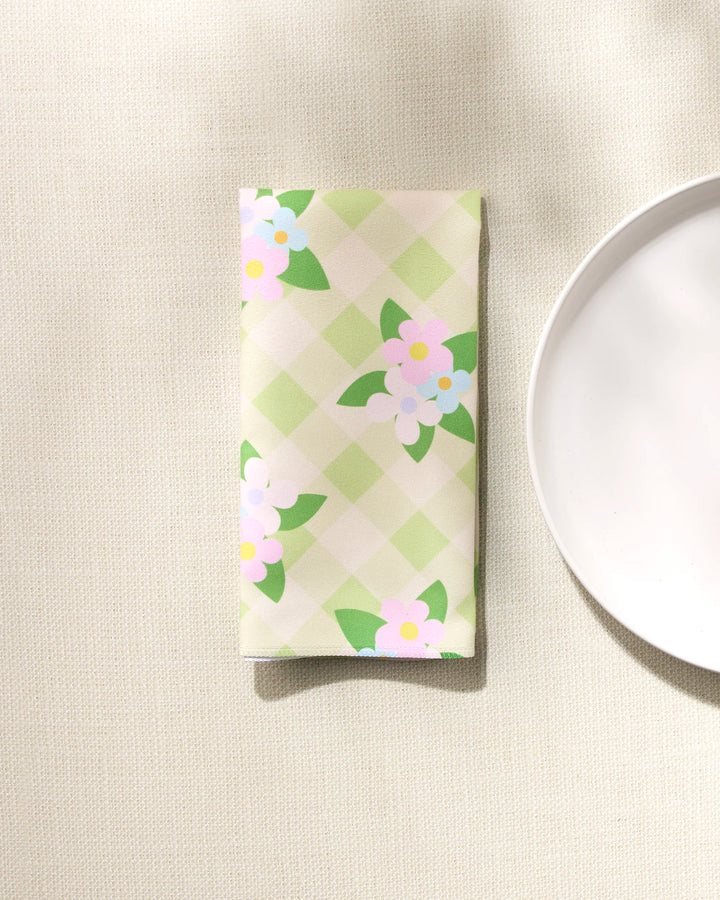 The Doting and Devoted folded dinner napkin set by GEOMETRY, made from recycled Geosuede fabric, showcases a green checkered pattern with pastel floral designs and rests beside a white plate on a textured surface.