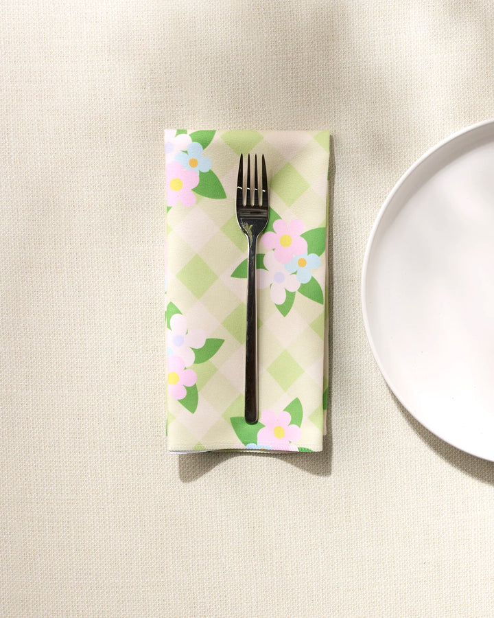 The GEOMETRY "Doting and Devoted" black fork is set on a pastel green and white checkered recycled dinner napkin with floral prints, placed beside a white plate on a light textured surface.