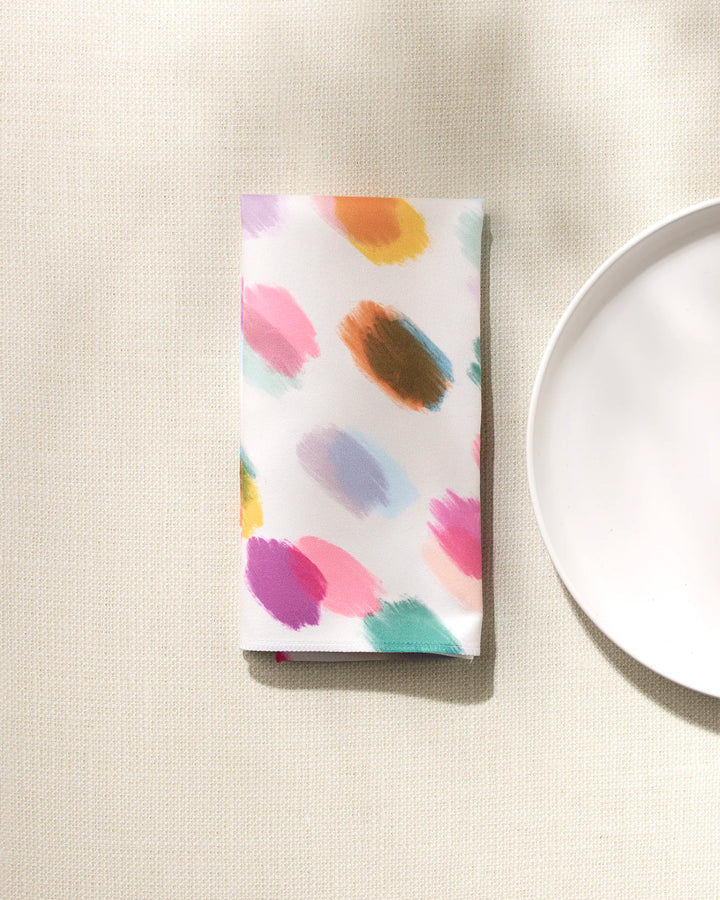 The GEOMETRY Downpour absorbent cloth napkin, featuring colorful brushstroke patterns, is folded on a textured beige surface beside the edge of a plain white plate.