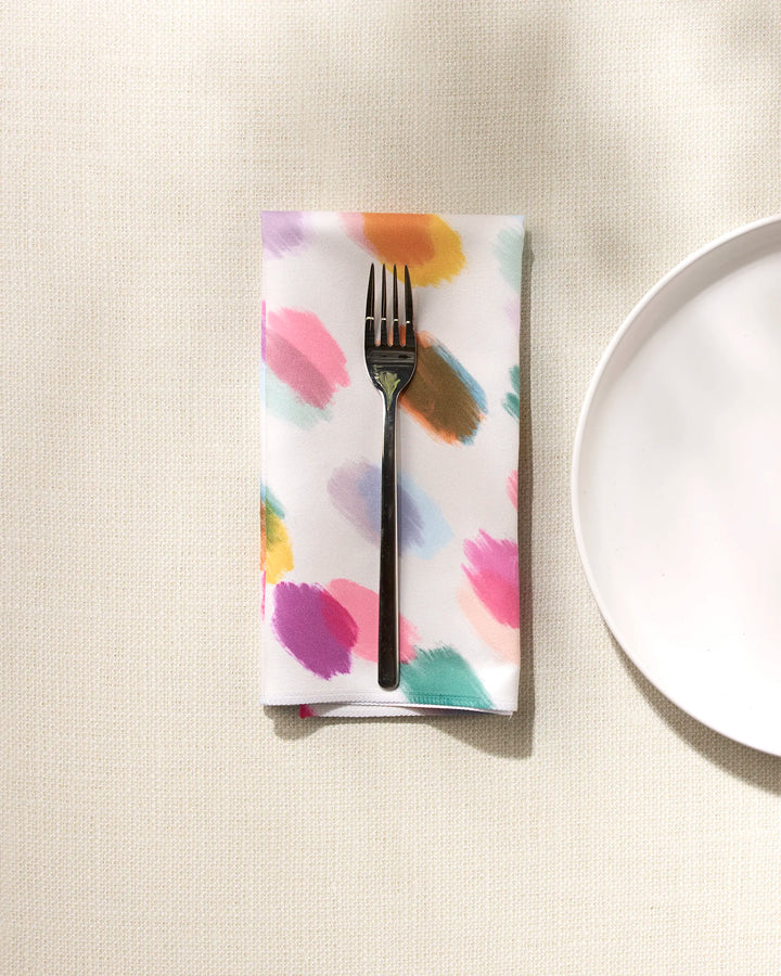 A black fork rests on GEOMETRY's Downpour absorbent cloth napkin, featuring colorful paint-like strokes, next to a plain white plate on a textured, light surface.