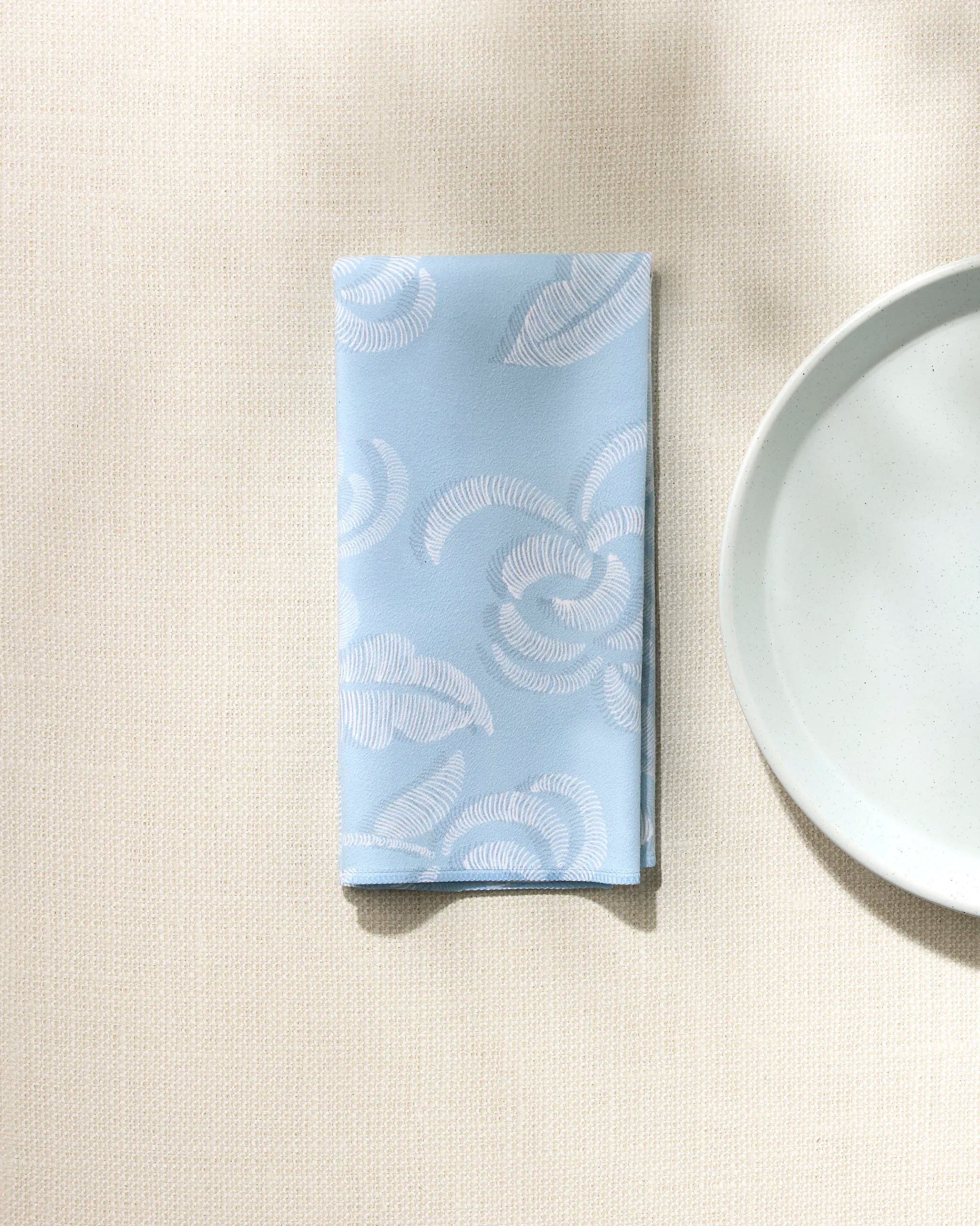 Edith's Embroidery by GEOMETRY is a light blue reusable napkin with a white swirl pattern, elegantly folded beside a white plate on a textured cream tablecloth—an ideal addition to any sophisticated dinner napkin set.