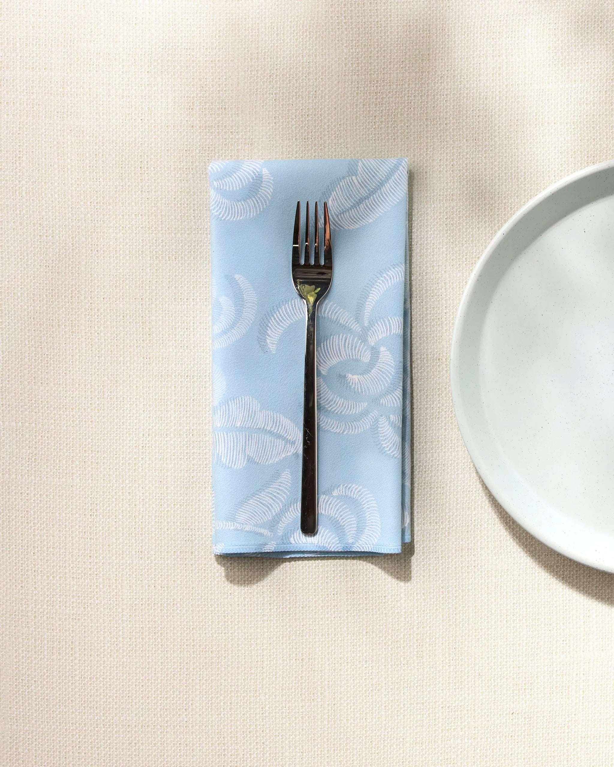The GEOMETRY Edith's Embroidery napkin in light blue with a white pattern is folded on a beige textured surface, topped with a silver fork and set beside a white ceramic plate.