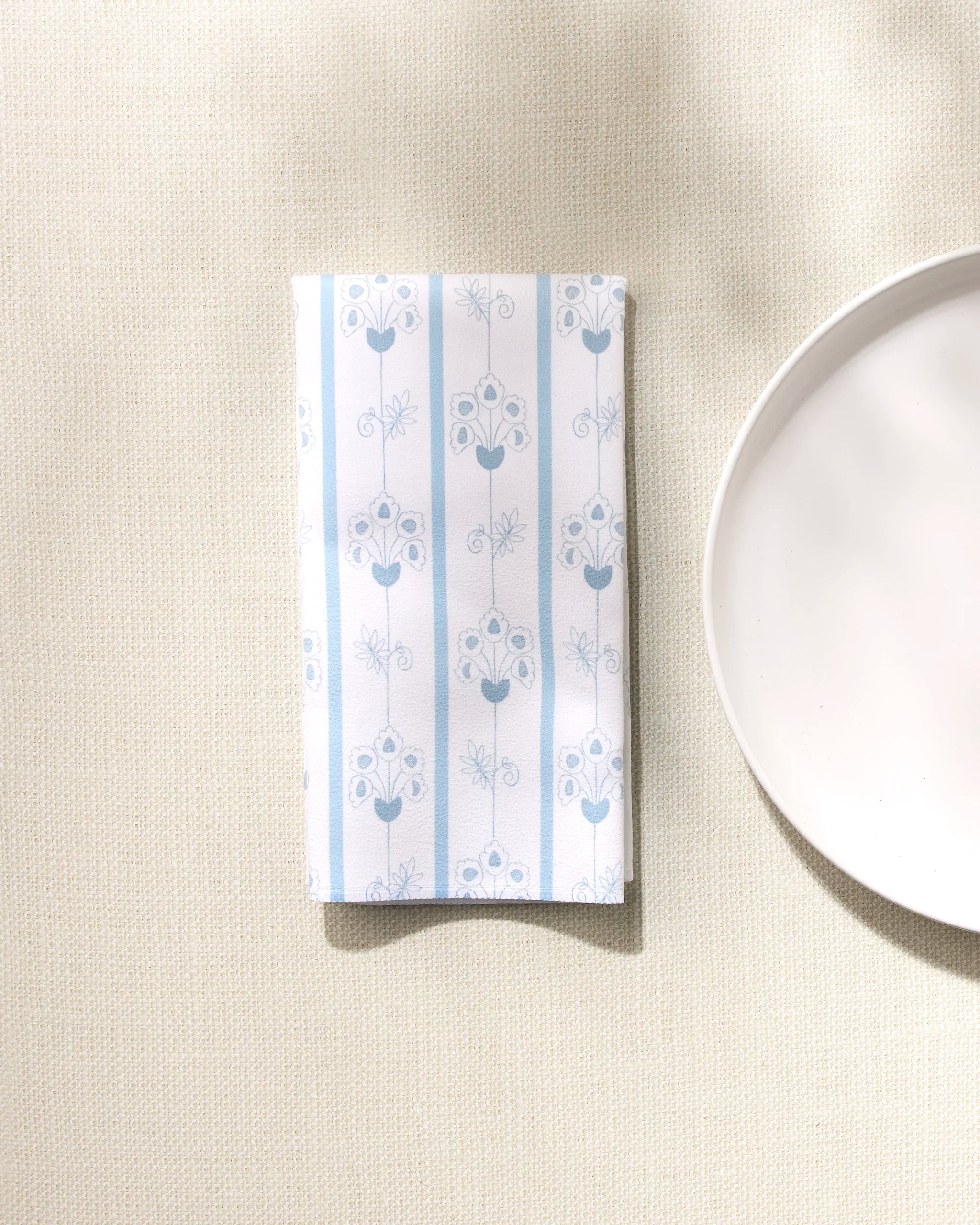 The Edith's English Cottage reusable napkin by GEOMETRY, featuring a light blue floral and striped design, rests on a textured beige surface beside a plain white plate with soft shadows across the scene.