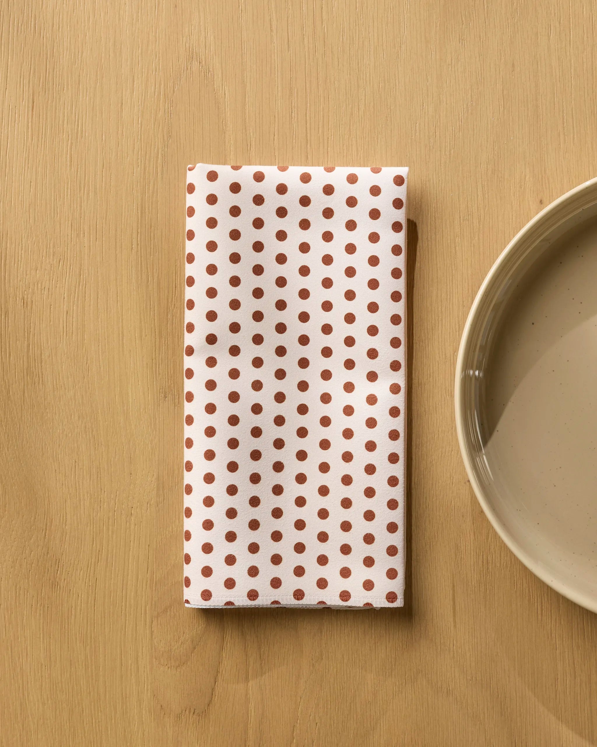 The Geometry Espresso Polka reusable napkin, featuring small brown polka dots on white, is folded on a light wooden surface beside a beige ceramic plate.