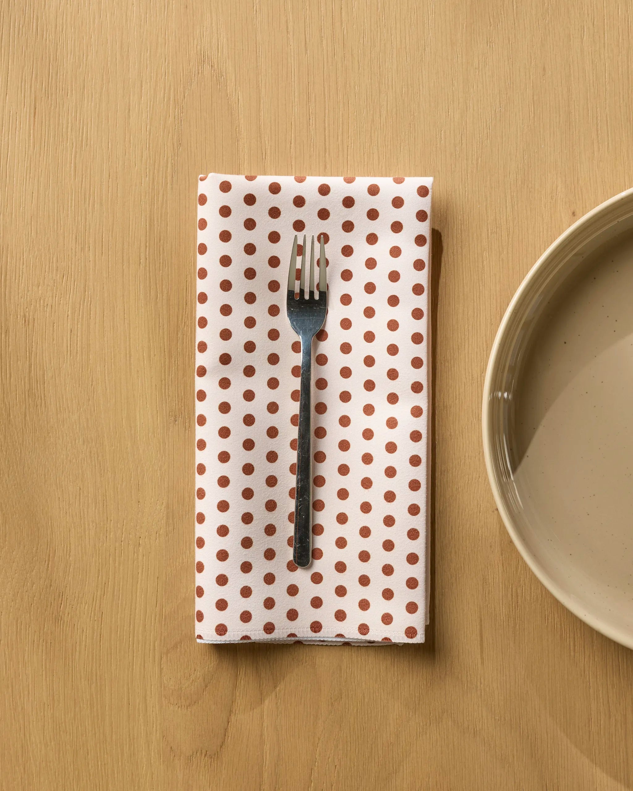 A metal fork sits on Geometry’s Espresso Polka reusable napkin—red polka dots on display—set beside a beige ceramic plate atop a light wooden table, making any meal more charming.