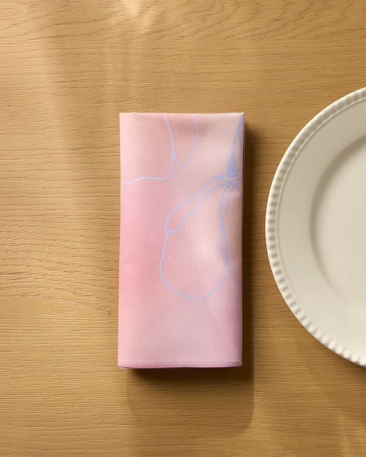 The Geometry Ethereal Bloom folded pink reusable dinner napkin, featuring a subtle floral pattern, rests on a light wooden table beside a white plate with a decorative rim.