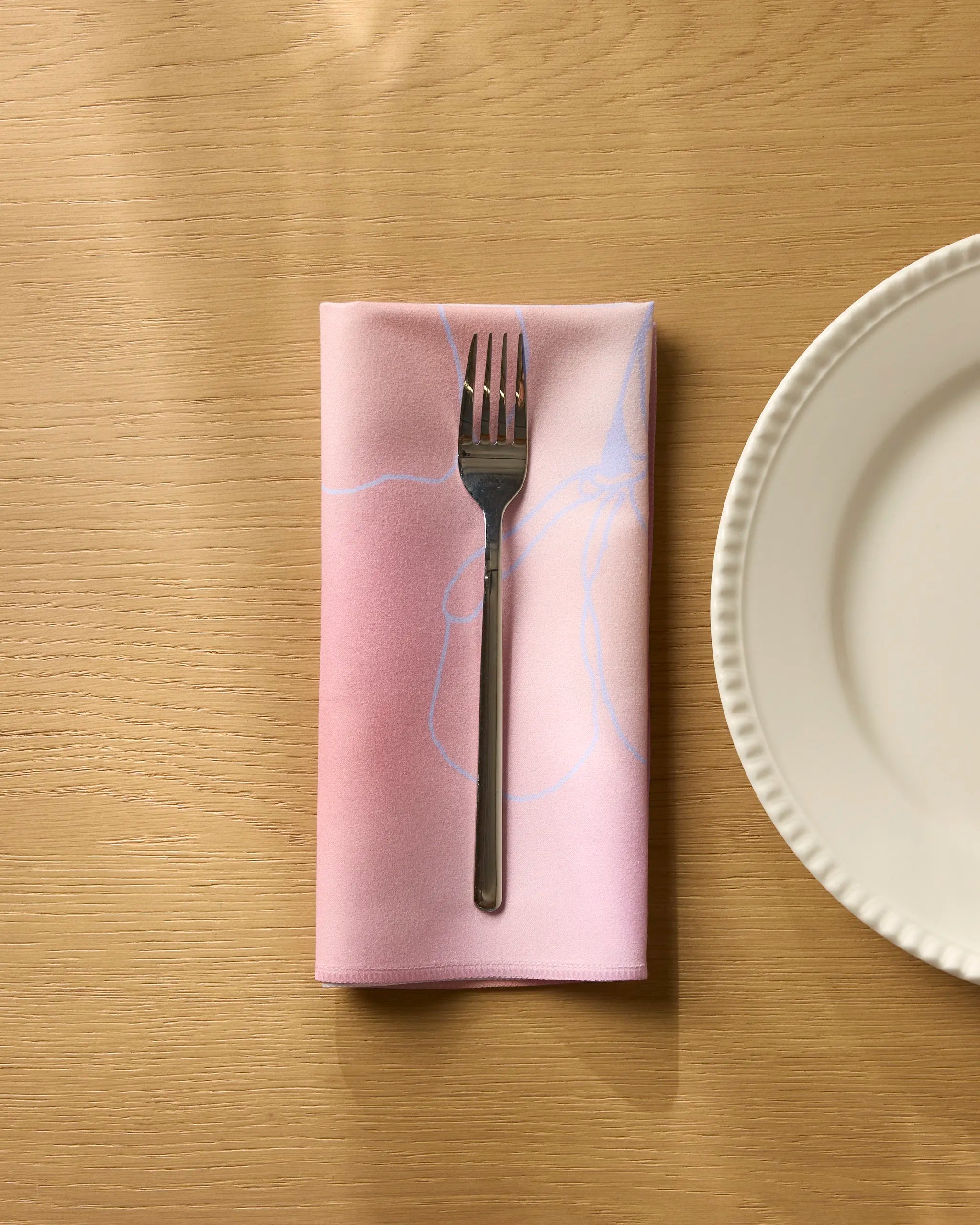 A silver fork rests on a folded Geometry Ethereal Bloom reusable napkin with subtle patterns, placed on a light wooden table beside a white ceramic plate with a scalloped edge.