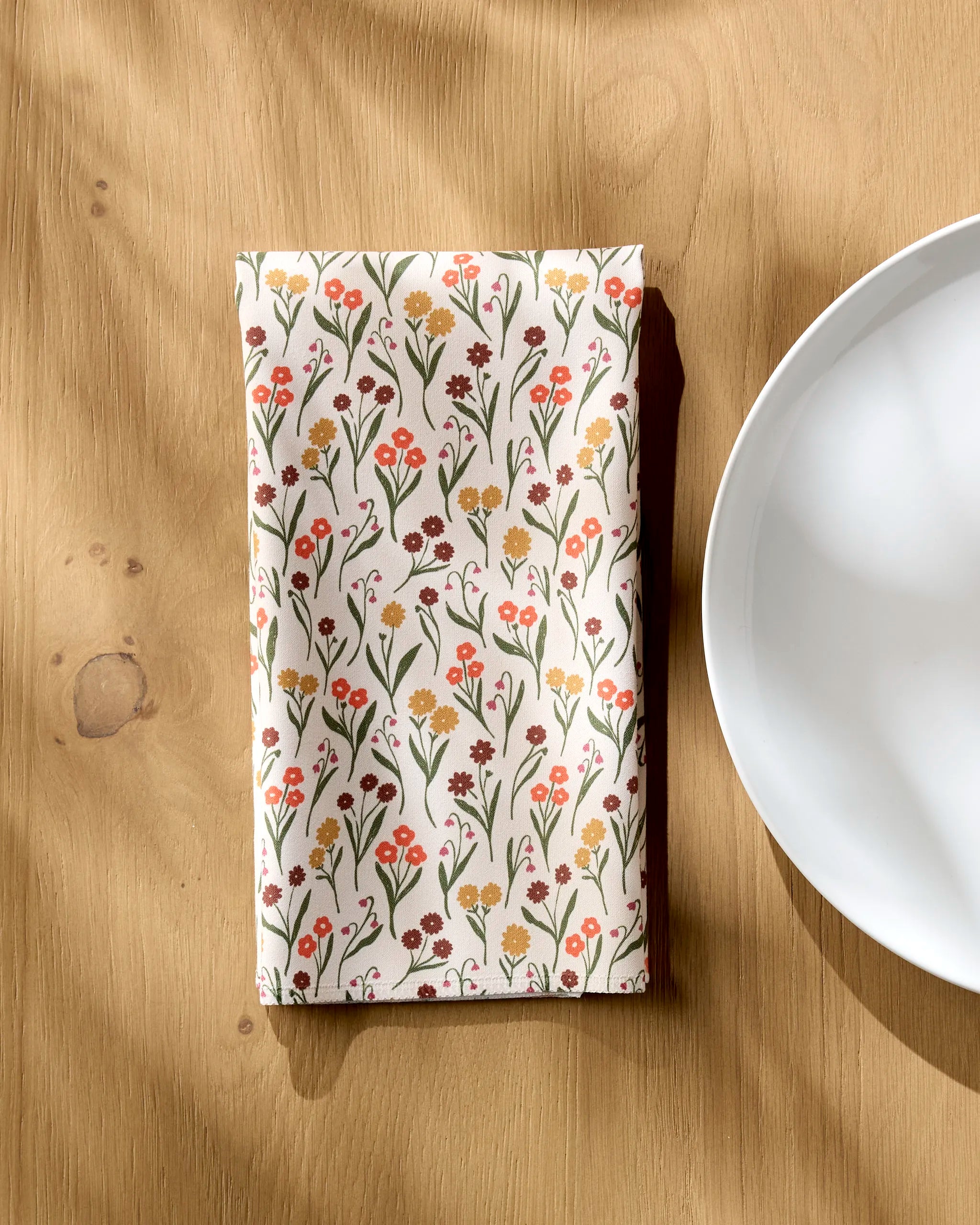 A folded Kayla Ann Fall Blossom reusable dinner napkin with a colorful floral pattern rests on a wooden table beside a white plate, sunlight highlighting the set’s eco-friendly elegance.