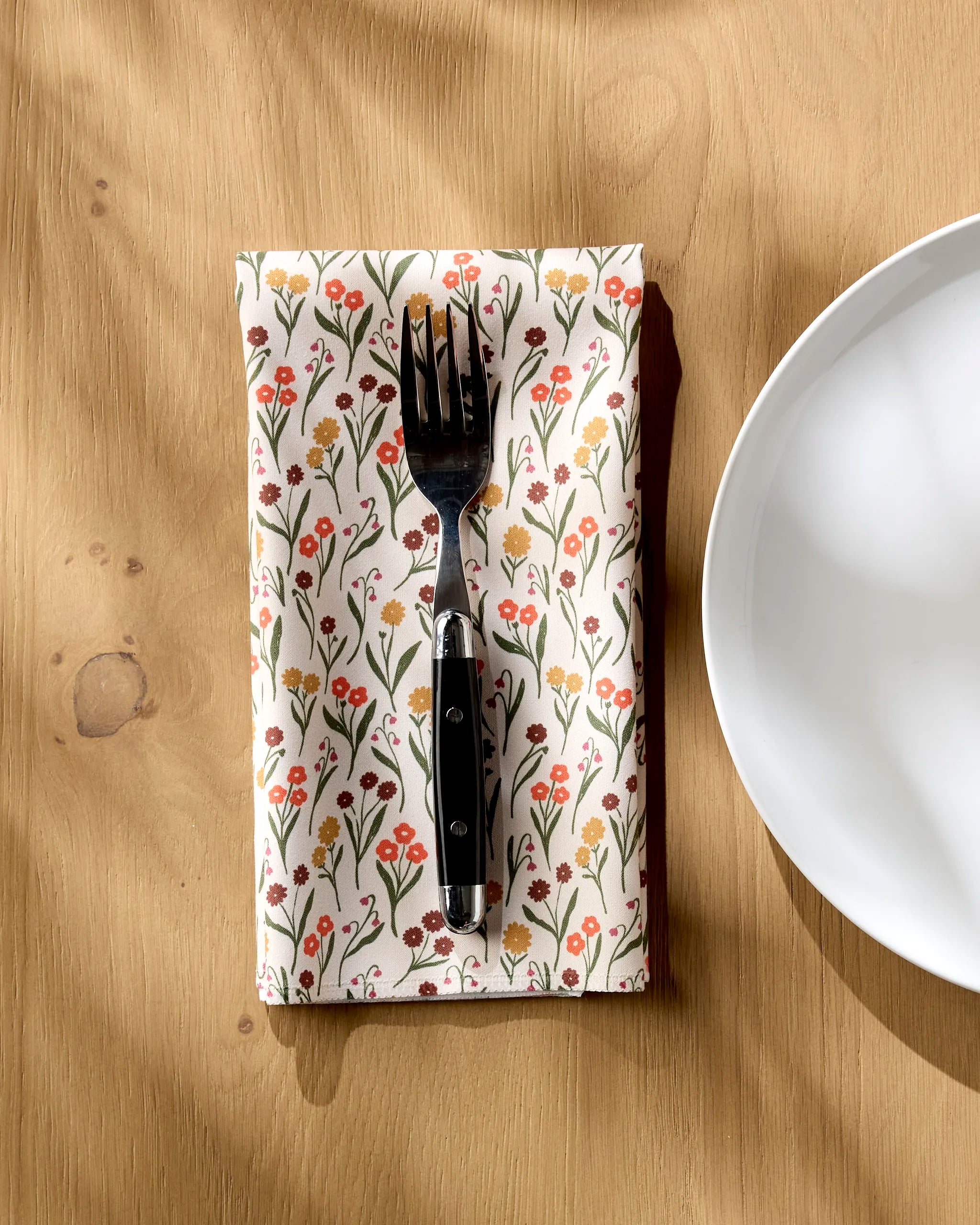 A Kayla Ann "Fall Blossom" black-handled fork rests on a folded, floral-patterned eco napkin by a white plate on a light wood table, soft sunlight casting gentle shadows.