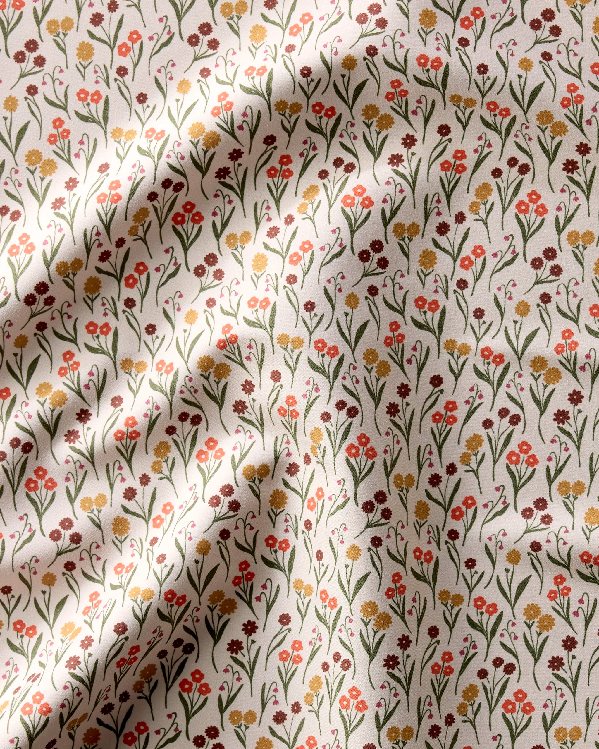 The Kayla Ann Fall Blossom set features reusable dinner napkins with a small floral pattern—red, yellow, and brown flowers with green stems on a light background. Soft shadows create gentle waves and highlight the recycled fabric’s texture.