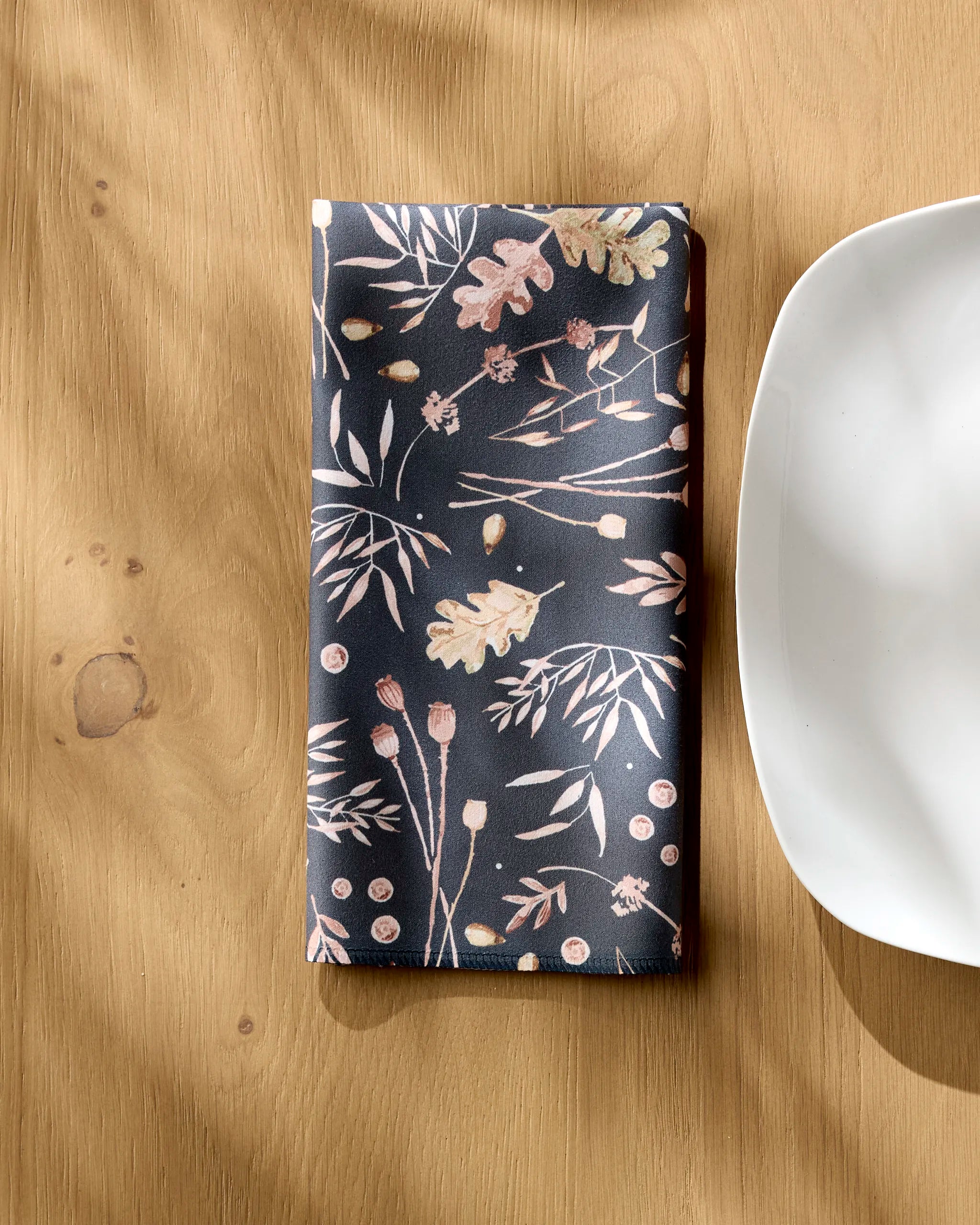 The Fallen Oak reusable cloth napkin by Katie O'Shea, featuring a botanical pattern, is folded neatly on a wooden table beside a round white plate, with sunlight casting gentle shadows over the scene.