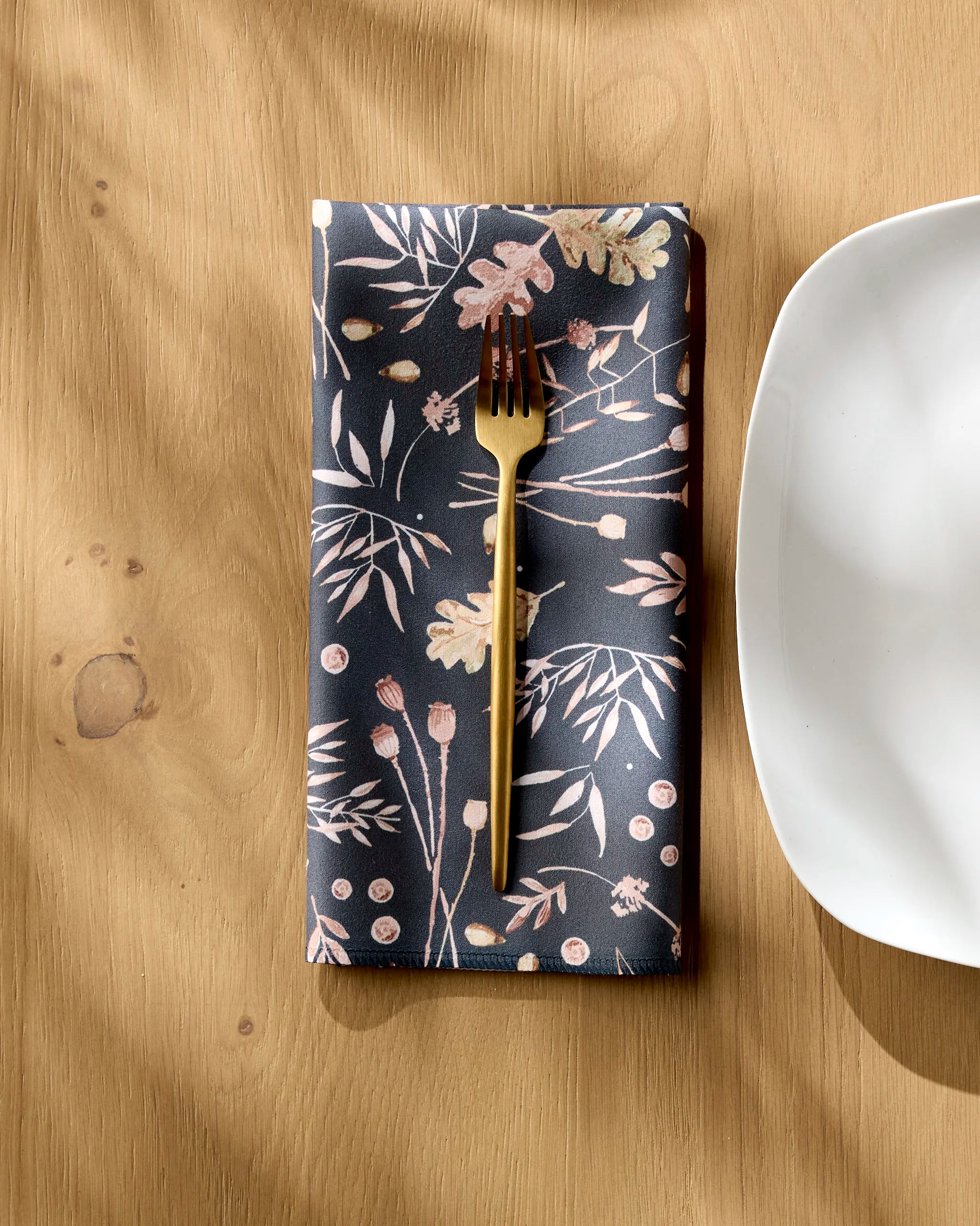 A gold fork rests on the Fallen Oak reusable cloth napkin by Katie O'Shea, featuring a botanical print and folded neatly on a light wooden table beside a white, rounded-edge plate.