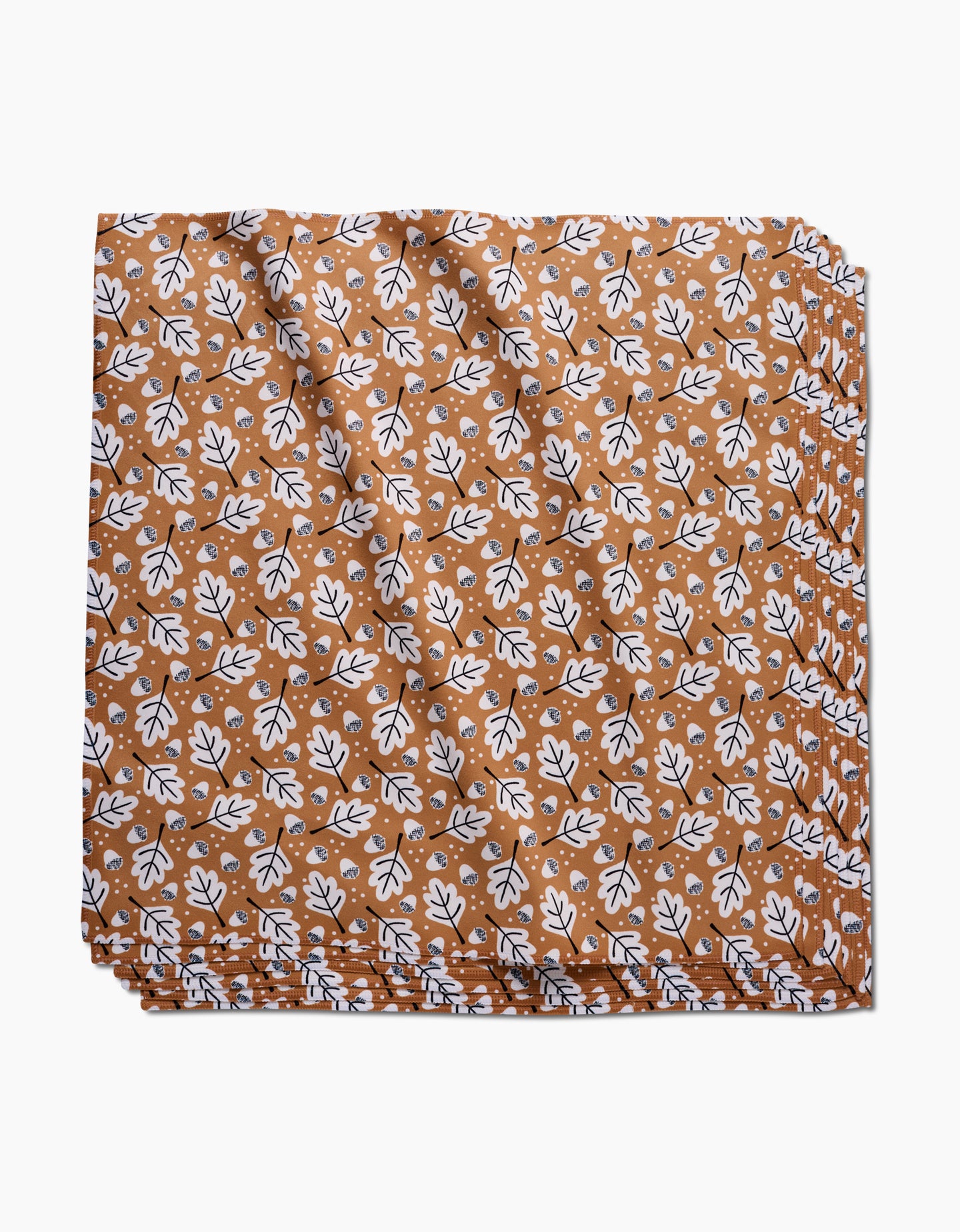 The Falling Acorns table linen by Charlie Rowan Designs features a brown background with a repeating white and black leaf pattern. This eco-friendly fabric is neatly folded and stacked, shown against a white backdrop.