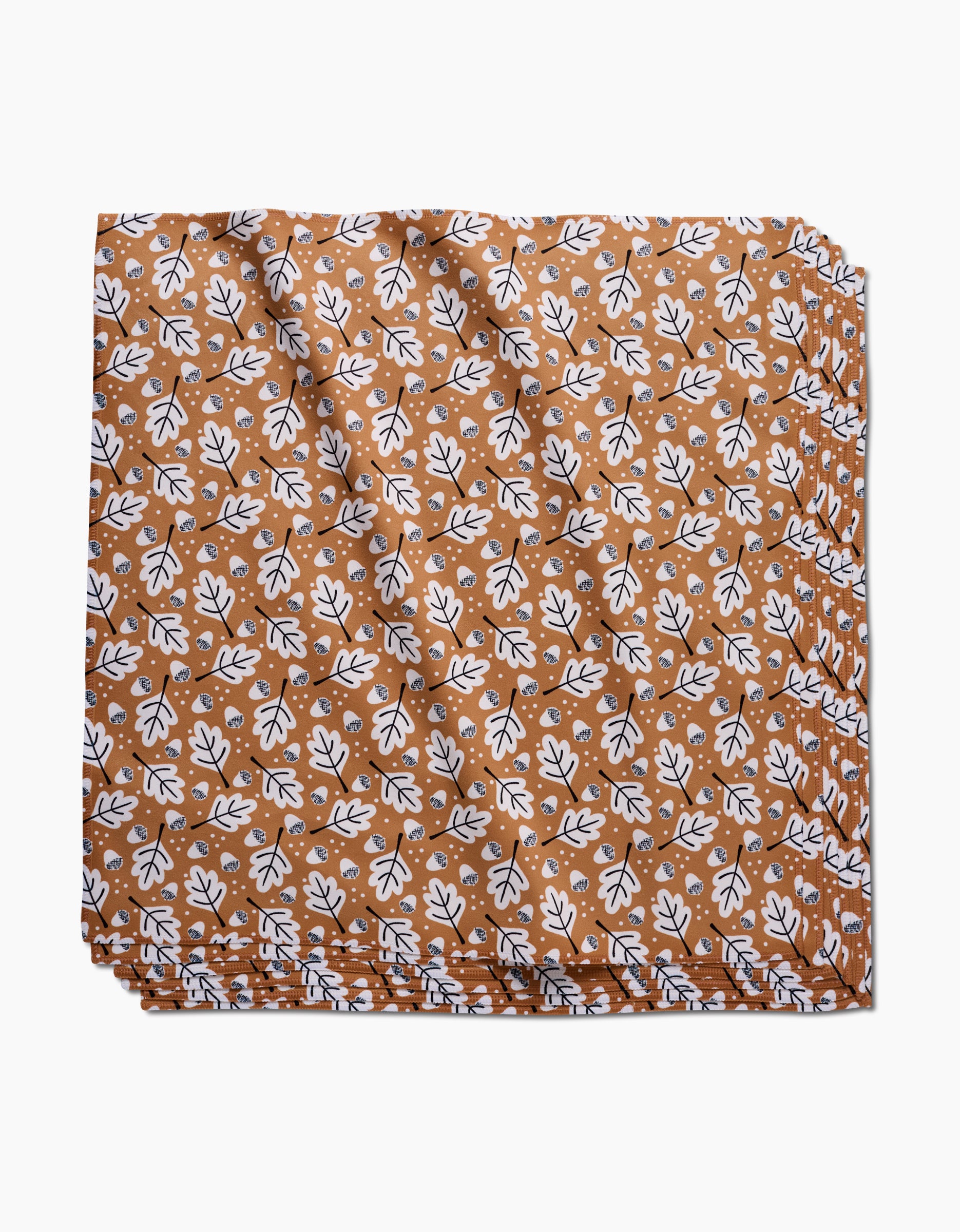 The Falling Acorns table linen by Charlie Rowan Designs features a brown background with a repeating white and black leaf pattern. This eco-friendly fabric is neatly folded and stacked, shown against a white backdrop.