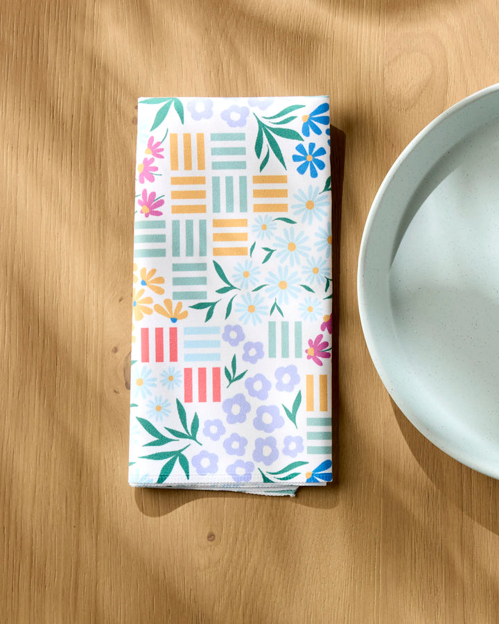A folded Charlie Rowan Designs "Floral Garden" eco-friendly napkin with vibrant floral and geometric patterns rests on a wooden table by a white plate, sunlight highlighting the sustainable choice of this Dinner Napkin Set.
