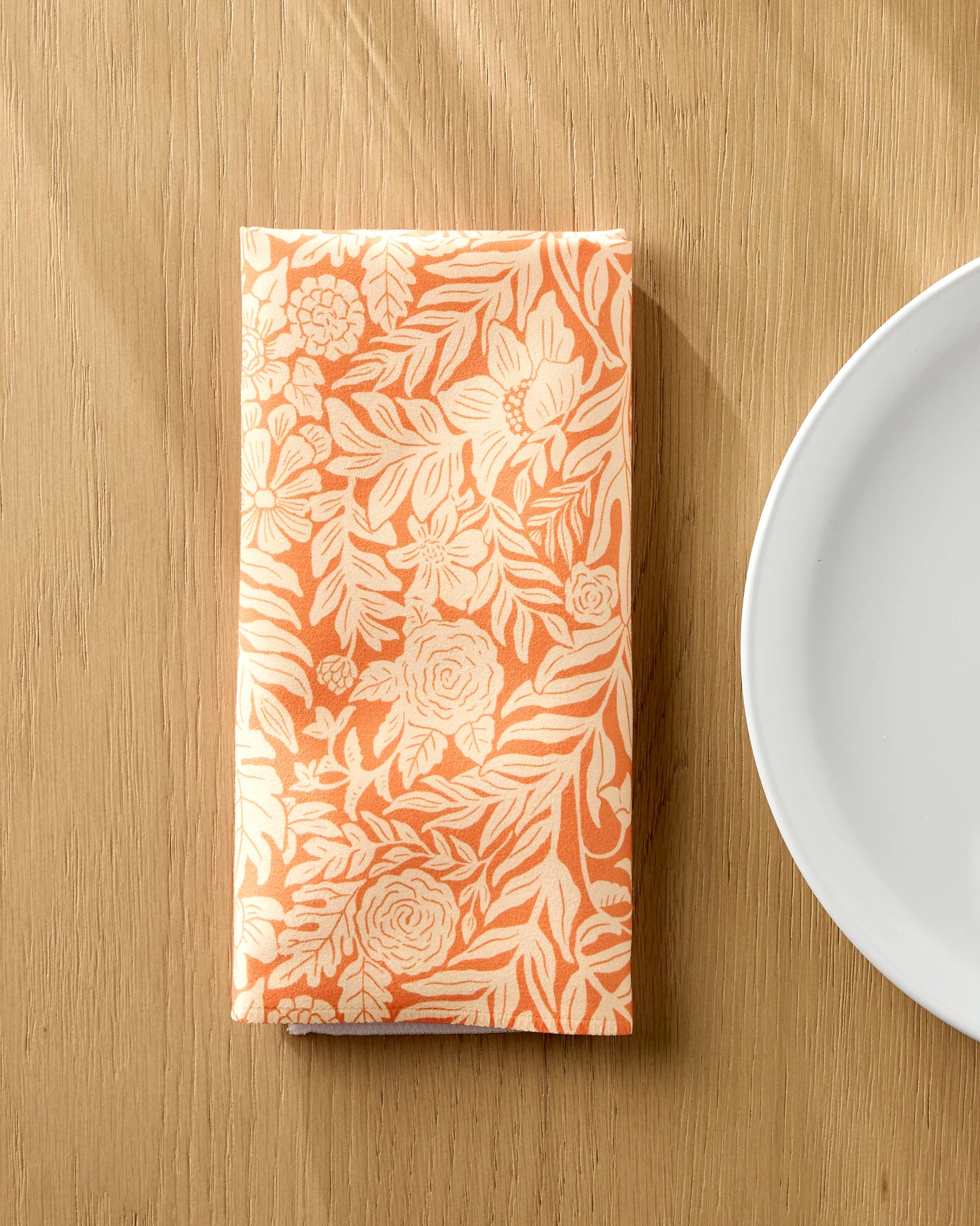 A folded orange Florence napkin by Katie O'Shea, featuring a cream floral pattern from the absorbent cloth napkins set, rests on a light wooden table beside a white plate.