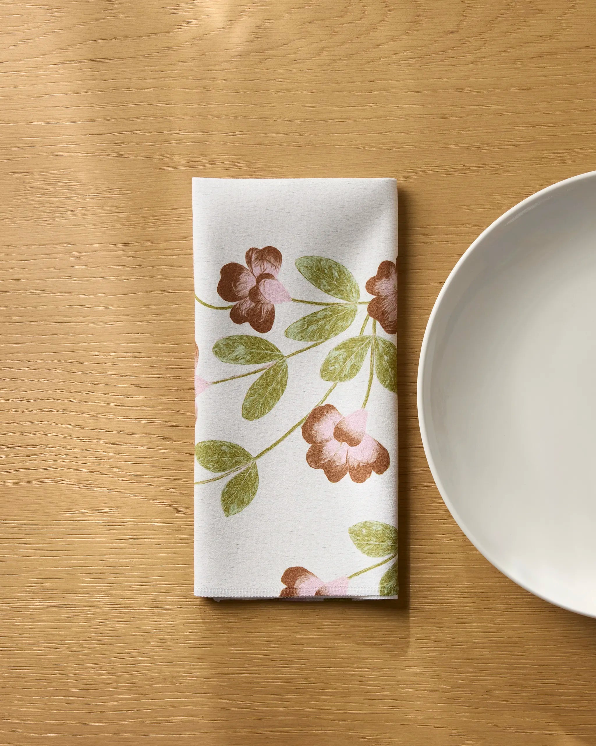 The Geometry Flower Tower napkin, folded and eco-friendly with a green and brown floral pattern, sits next to a white plate on a light wooden table.
