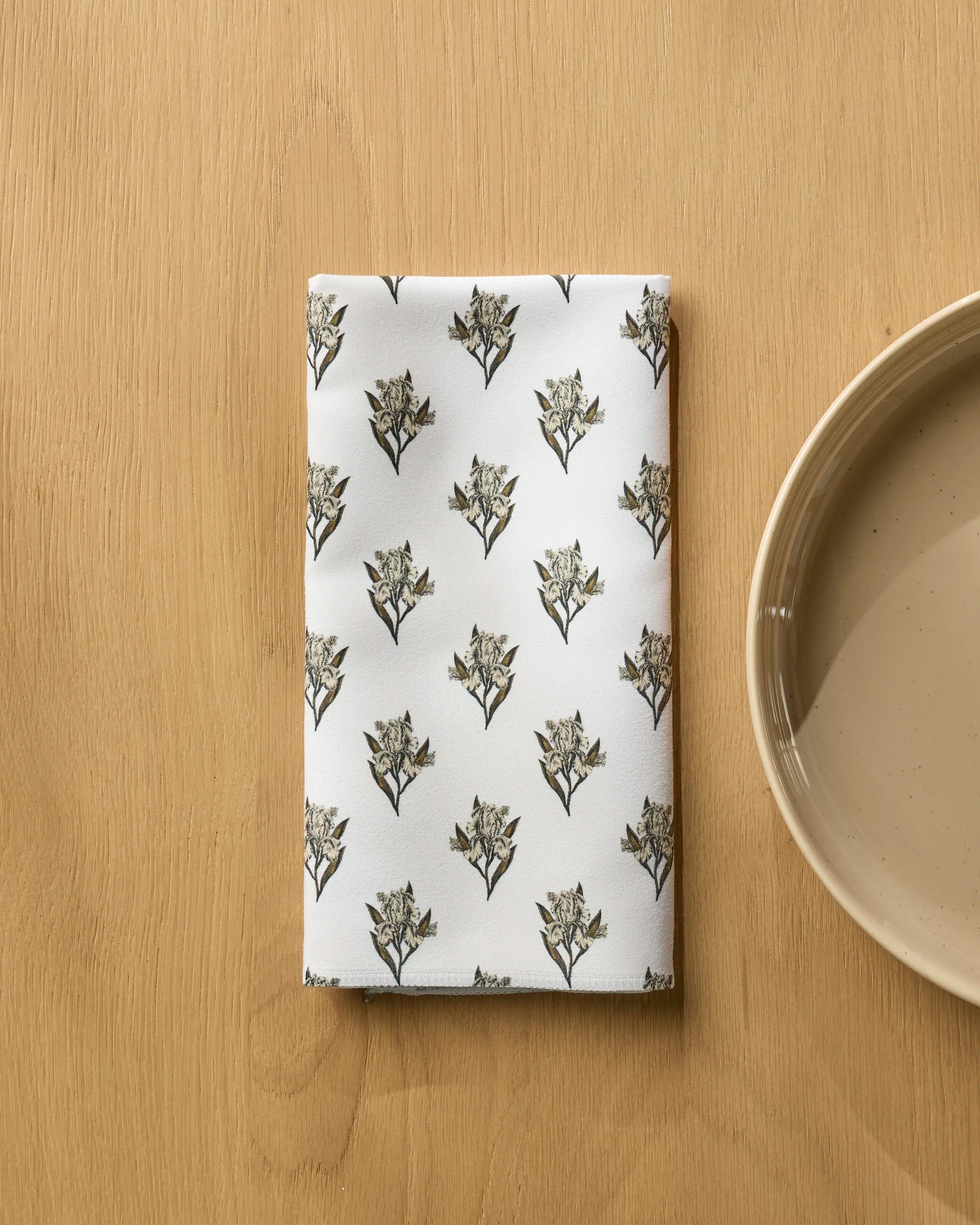 The Foraged recycled dinner napkin set by Geometry features a folded white napkin with a black and gray floral pattern on a light wooden surface beside a beige ceramic plate, bringing eco-friendly style to your table.