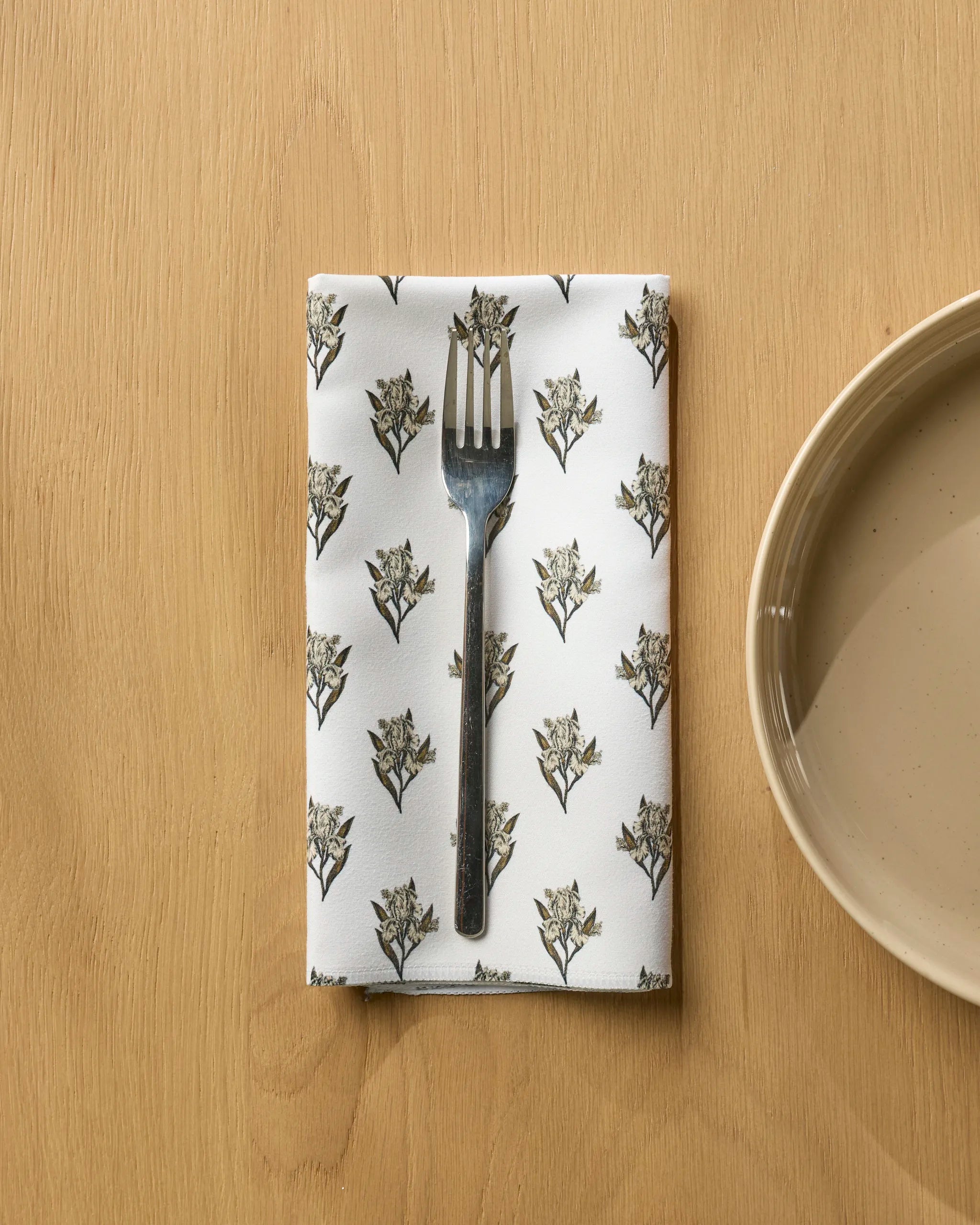 A Geometry "Foraged" metal fork rests on a white recycled napkin with a floral pattern, placed on a light wooden table beside a beige ceramic plate.
