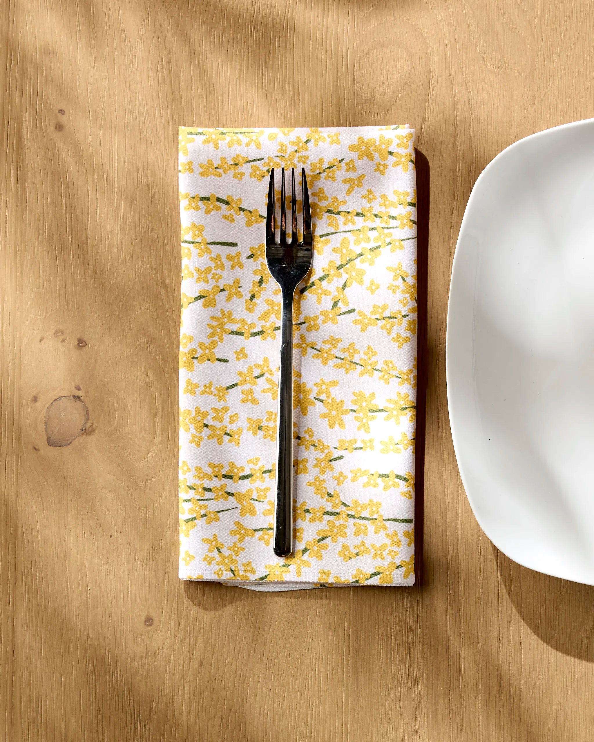 A metal fork rests on a Kayla Ann Forsythia Floral white reusable dinner napkin with a yellow floral pattern, placed beside a white, square-edged plate on a light wooden table. Sunlight creates soft shadows across the scene.