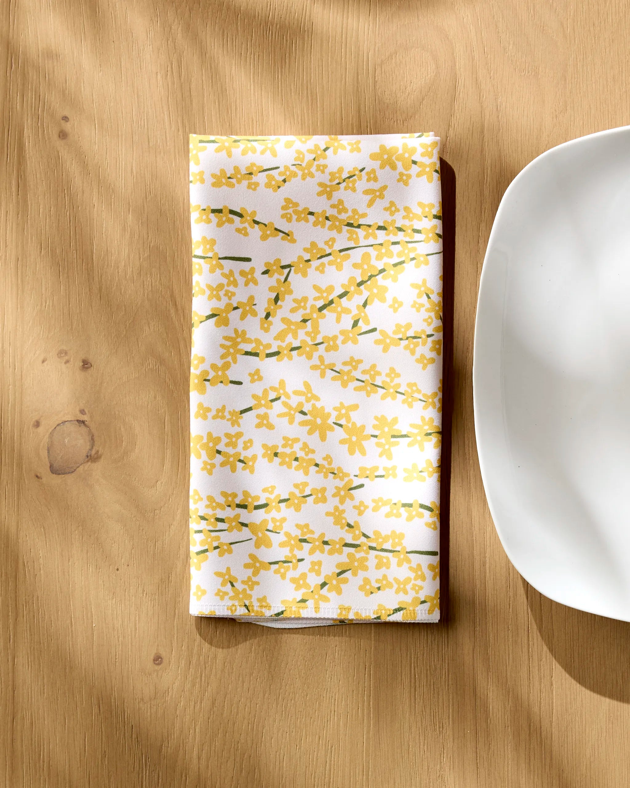 The Kayla Ann Forsythia Floral napkin, featuring a yellow floral pattern on white, is folded beside a square-edged plate on a light wooden table as sunlight casts gentle shadows.