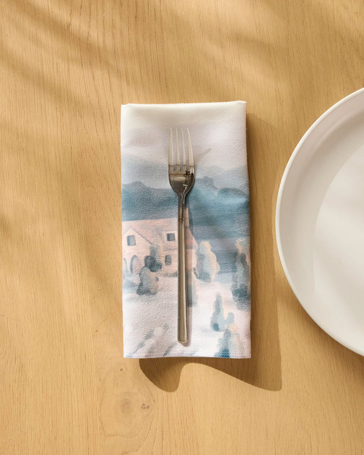 A fork rests on a folded Hannah Langford French Cottage recycled dinner napkin with a soft watercolor landscape design, placed next to a white plate on a light wooden table.