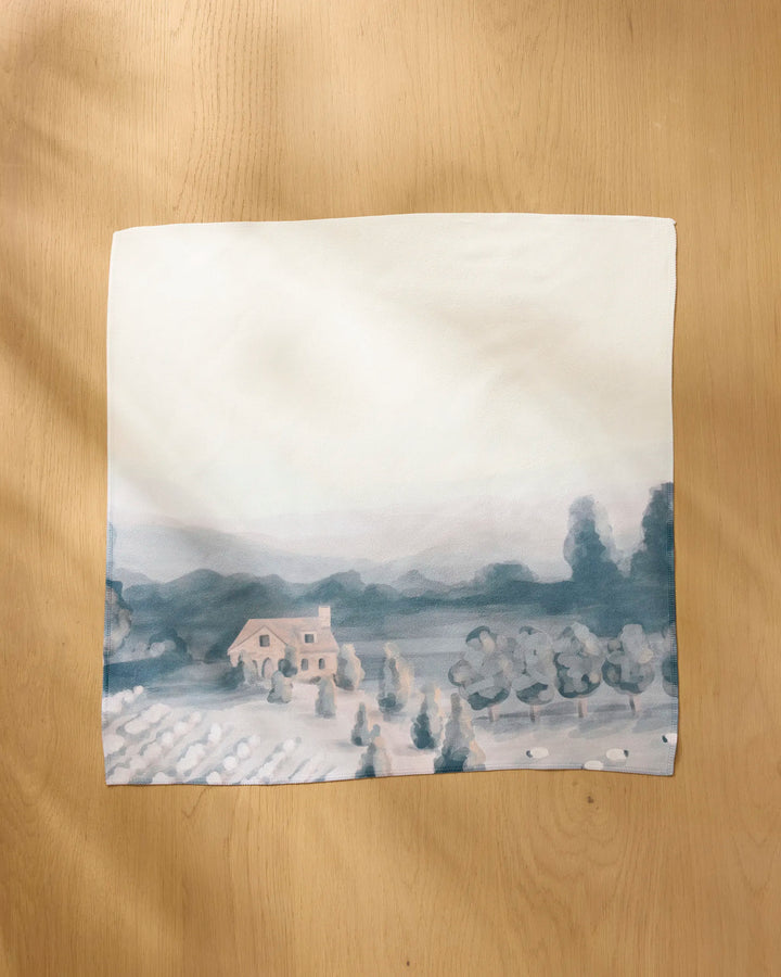 The "French Cottage" by Hannah Langford is a square painting on recycled dinner napkin paper, showing a house, trees, and fields in soft blue and beige tones, displayed on a wooden surface.