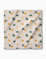 The Freshly Picked dinner napkin by Sara Mari features yellow lemons, green leaves, and lemon slices on light beige Geosuede made from recycled materials. Slight wrinkles in the fabric add a relaxed touch.