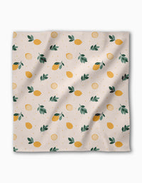 The Freshly Picked dinner napkin by Sara Mari features yellow lemons, green leaves, and lemon slices on light beige Geosuede made from recycled materials. Slight wrinkles in the fabric add a relaxed touch.