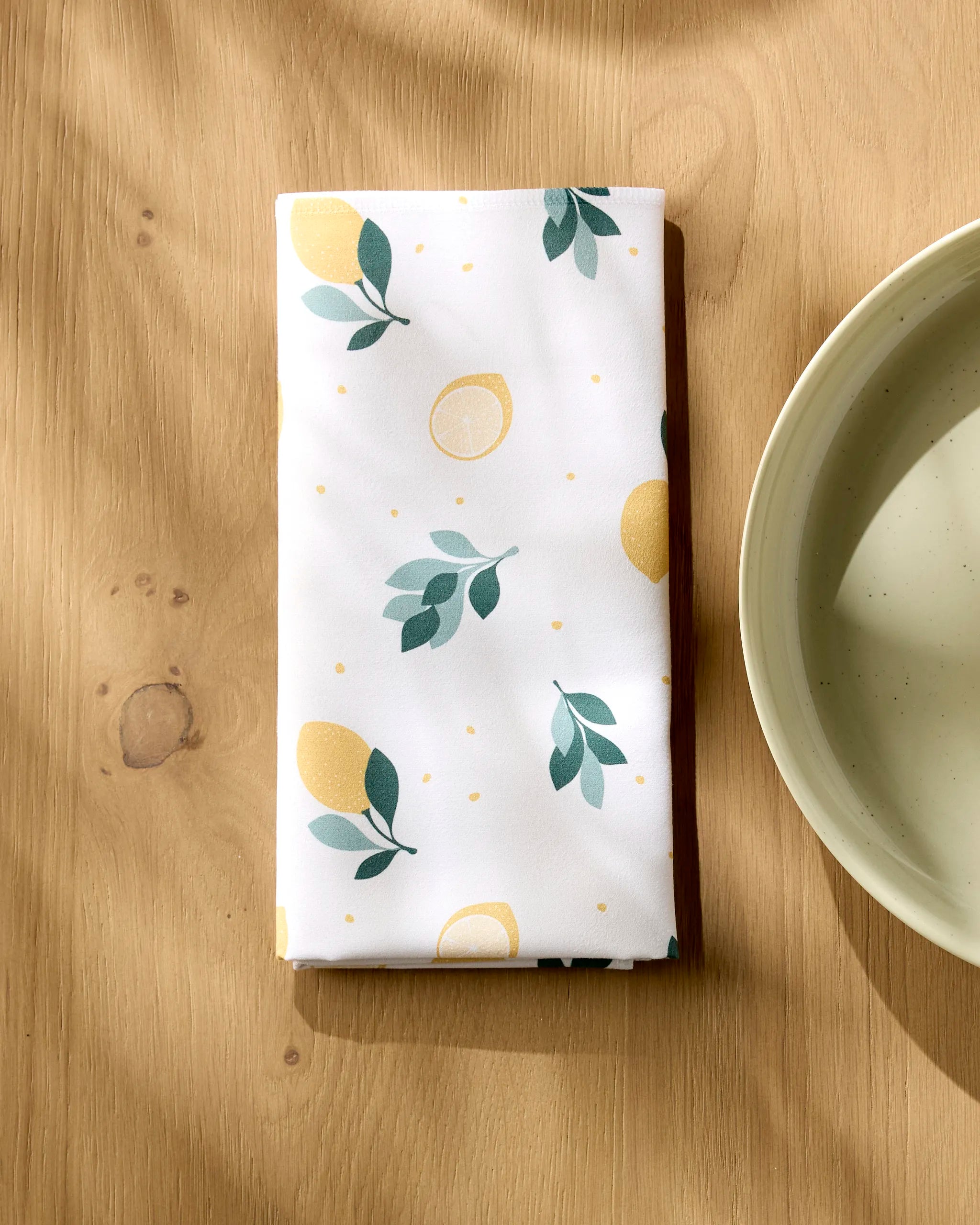 A Sara Mari "Freshly Picked" white absorbent dinner napkin with a lemon and leaf print is neatly folded on a light wooden table beside a ceramic bowl, with sunlight casting soft shadows.