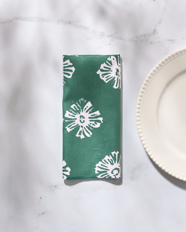 A Freya washable green cloth napkin by Geometry, featuring a white floral pattern, is placed on a white marble surface beside a scalloped-edge white ceramic plate.