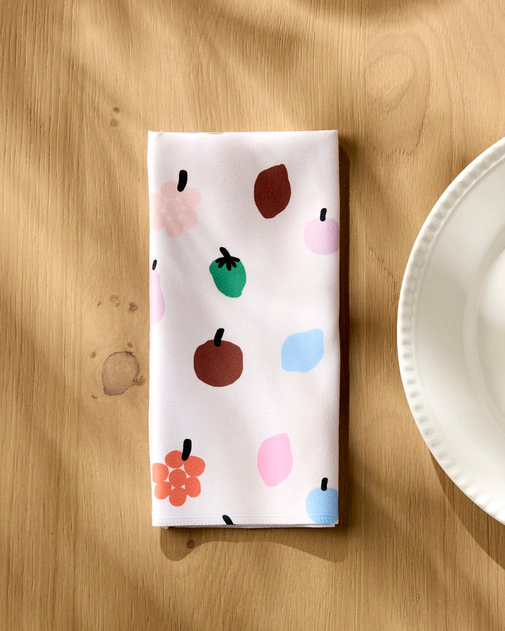 The Jennifer Bouron Fruit Basket napkin, featuring colorful abstract fruit and flower designs on white fabric, is folded on a light wooden table beside a scalloped white plate as sunlight casts gentle shadows.