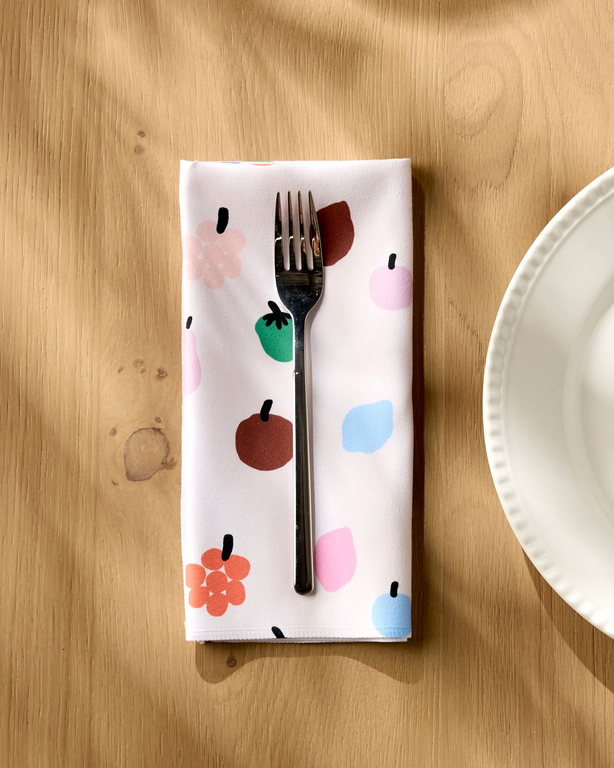 A silver fork rests on a white "Fruit Basket" dinner napkin by Jennifer Bouron, featuring colorful fruit and flower prints, placed on a light wooden table next to a white plate.