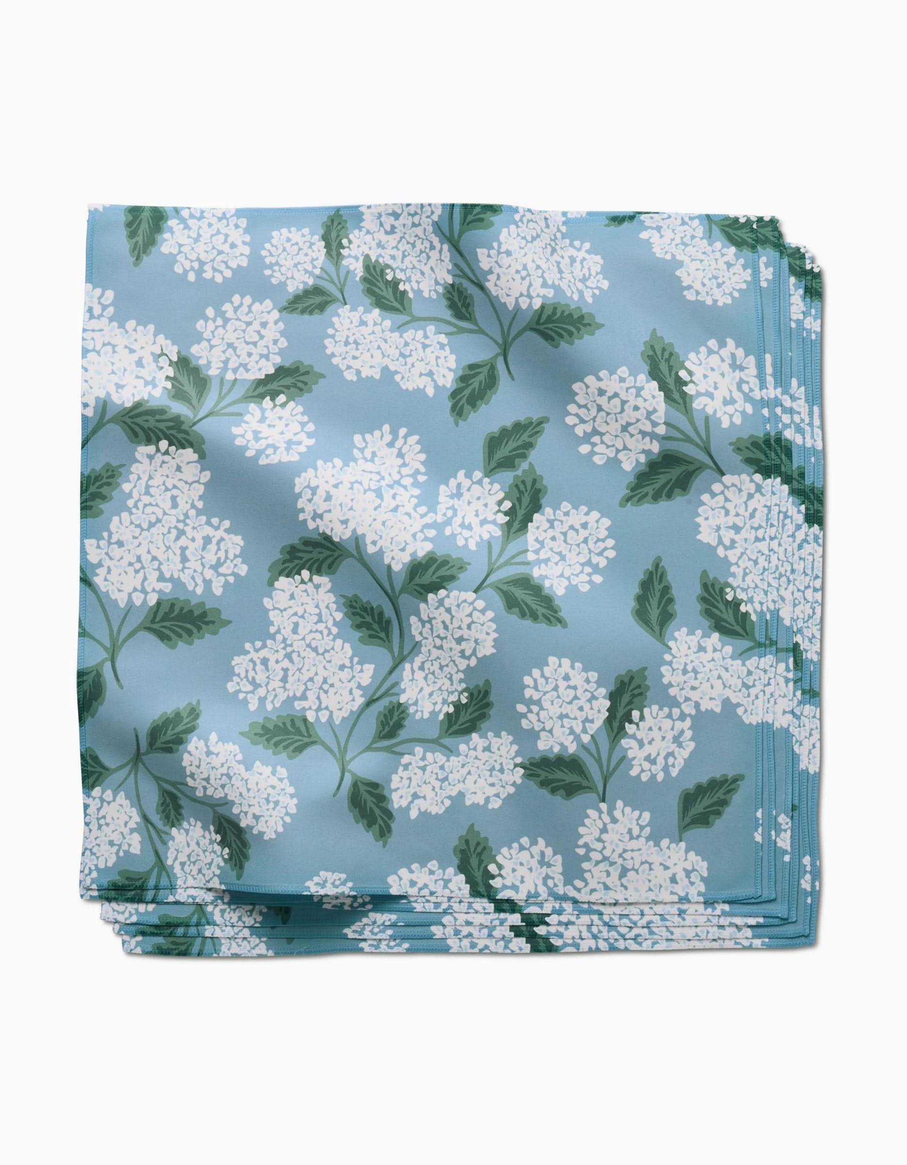 The GEOMETRY Garden Of Edith is a folded blue dinner napkin with white floral clusters and green leaves, crafted from soft Geosuede fabric made from recycled materials.