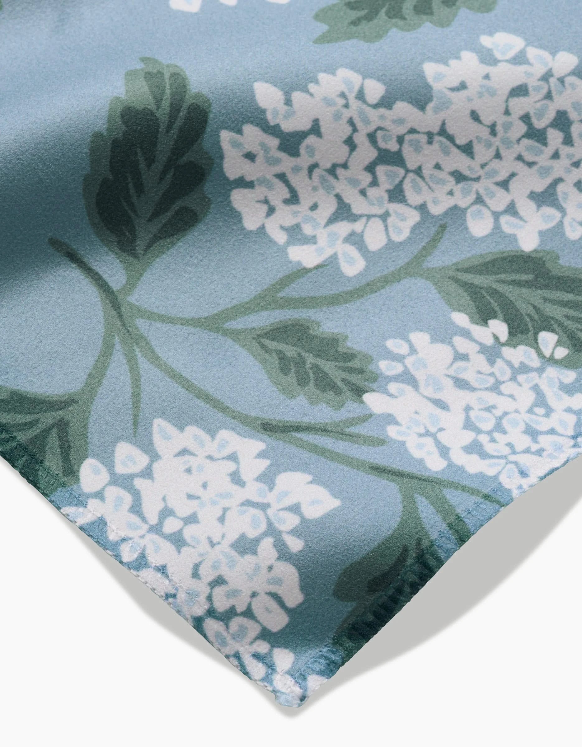 A close-up of GEOMETRY’s Garden Of Edith Geosuede fabric, light blue with clusters of white flowers and large green leaves, ideal for Dinner Napkins and shown folded at one corner.
