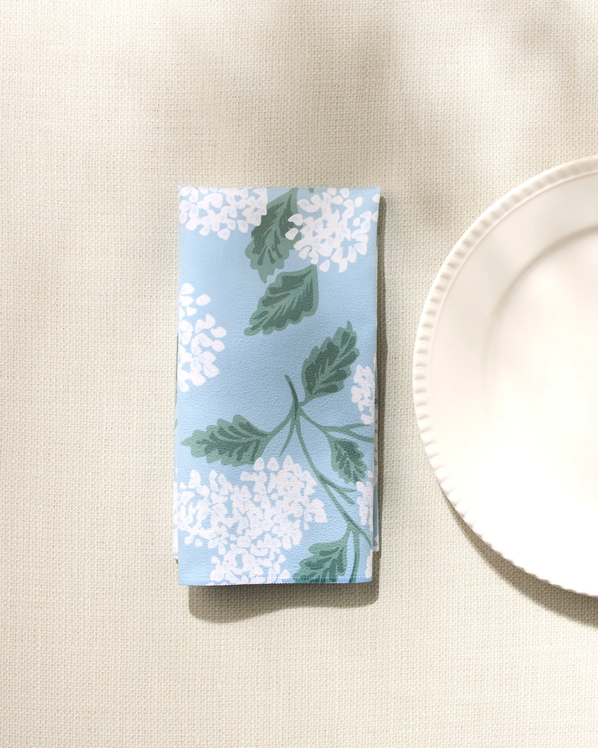 The GEOMETRY 'Garden Of Edith' reusable napkin, featuring a blue background with white florals and green leaves, is shown folded on a beige textured surface beside a white scalloped-edge plate—an elegant dinner napkin set.