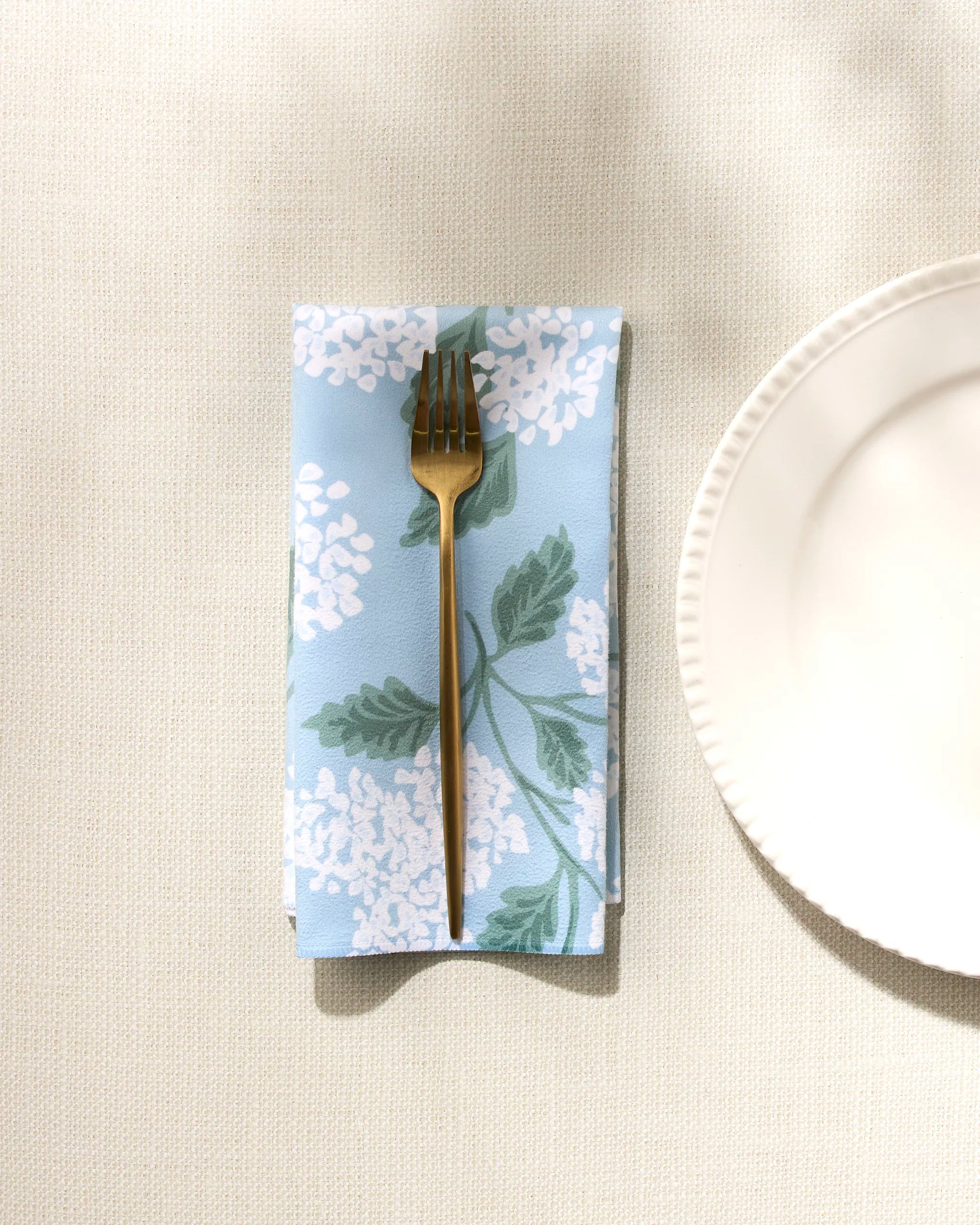 The GEOMETRY "Garden Of Edith" reusable blue napkin with white floral patterns rests under a gold fork beside a white plate on a textured light tablecloth.
